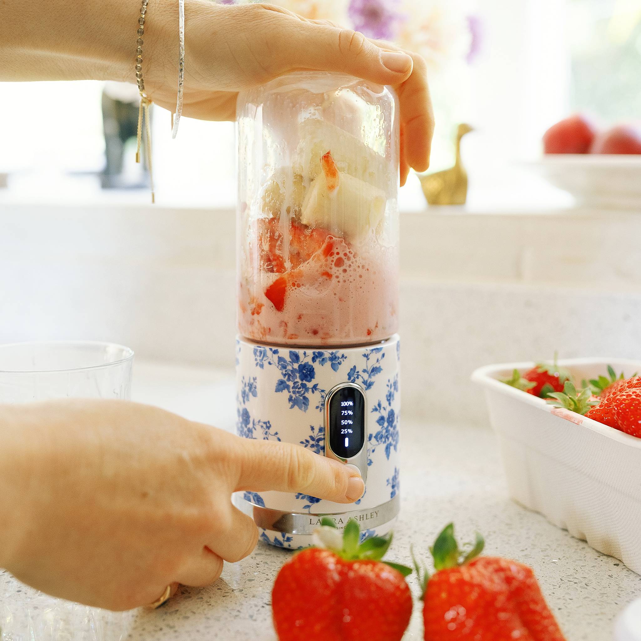 Eine Person benutzt einen Mixer mit Blumenmuster, um Erdbeeren und Joghurt zu mischen. Eine Schüssel mit Erdbeeren und andere Küchenutensilien sind im Hintergrund zu sehen.