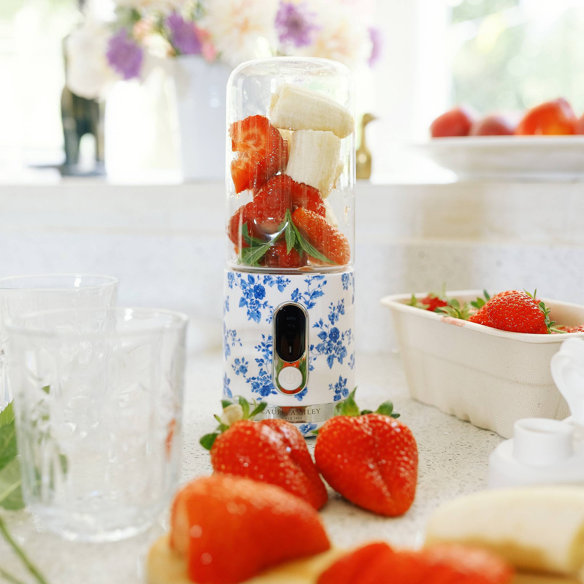 Ein Mixer gefüllt mit Erdbeeren und Bananen auf einer Küchenarbeitsplatte, umgeben von frischen Erdbeeren und Gläsern, mit Blumen im Hintergrund.