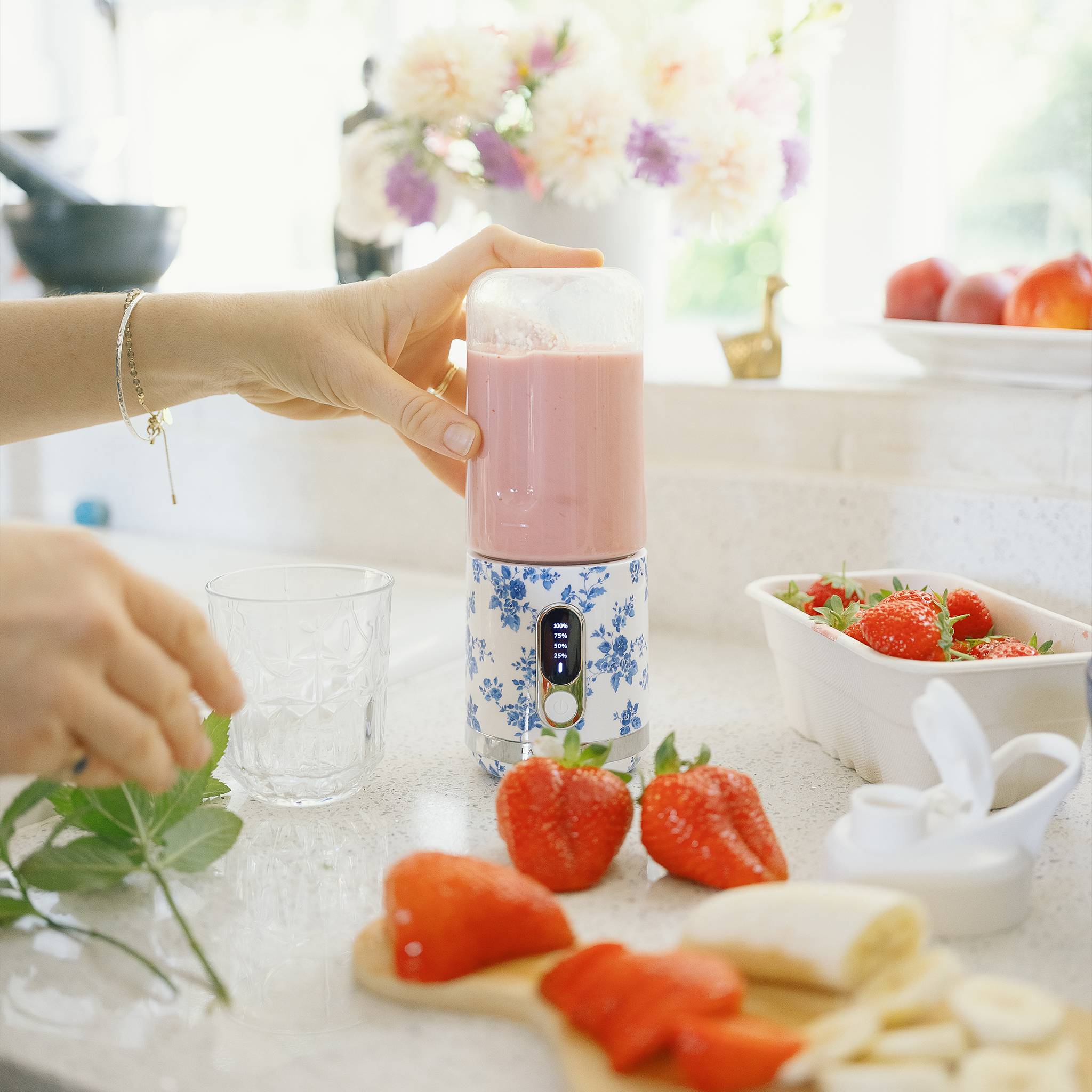 Eine Person bereitet einen Smoothie in einem blumigen Mixer zu, umgeben von Erdbeeren und Bananen auf einer Küchentheke mit einem Glas in der Nähe.