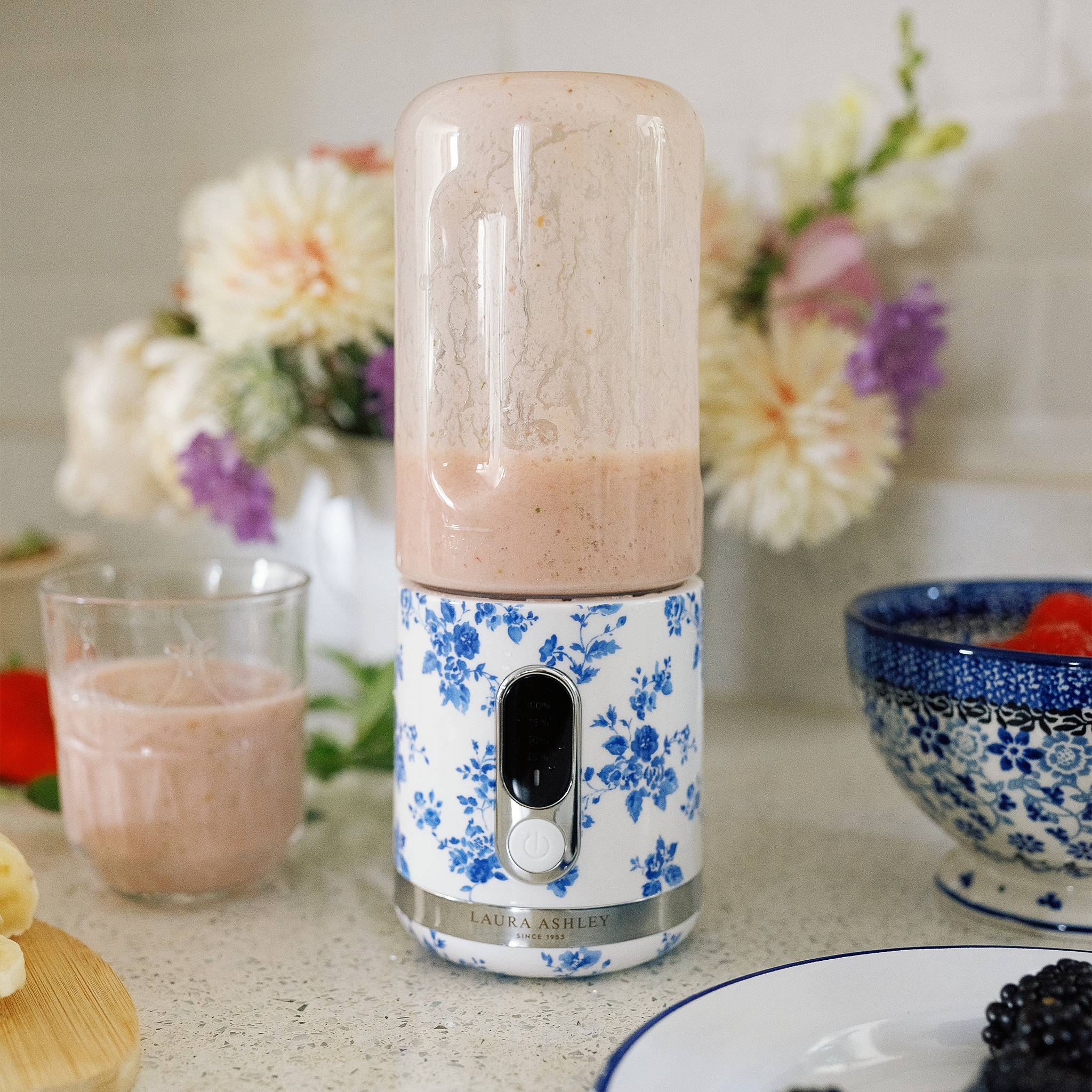 Ein Mixer mit Blumenmuster ist mit einem rosa Smoothie gefüllt, auf einer Küchenarbeitsplatte neben einem Glas des gleichen Getränks platziert, umgeben von Blumen und Obst.