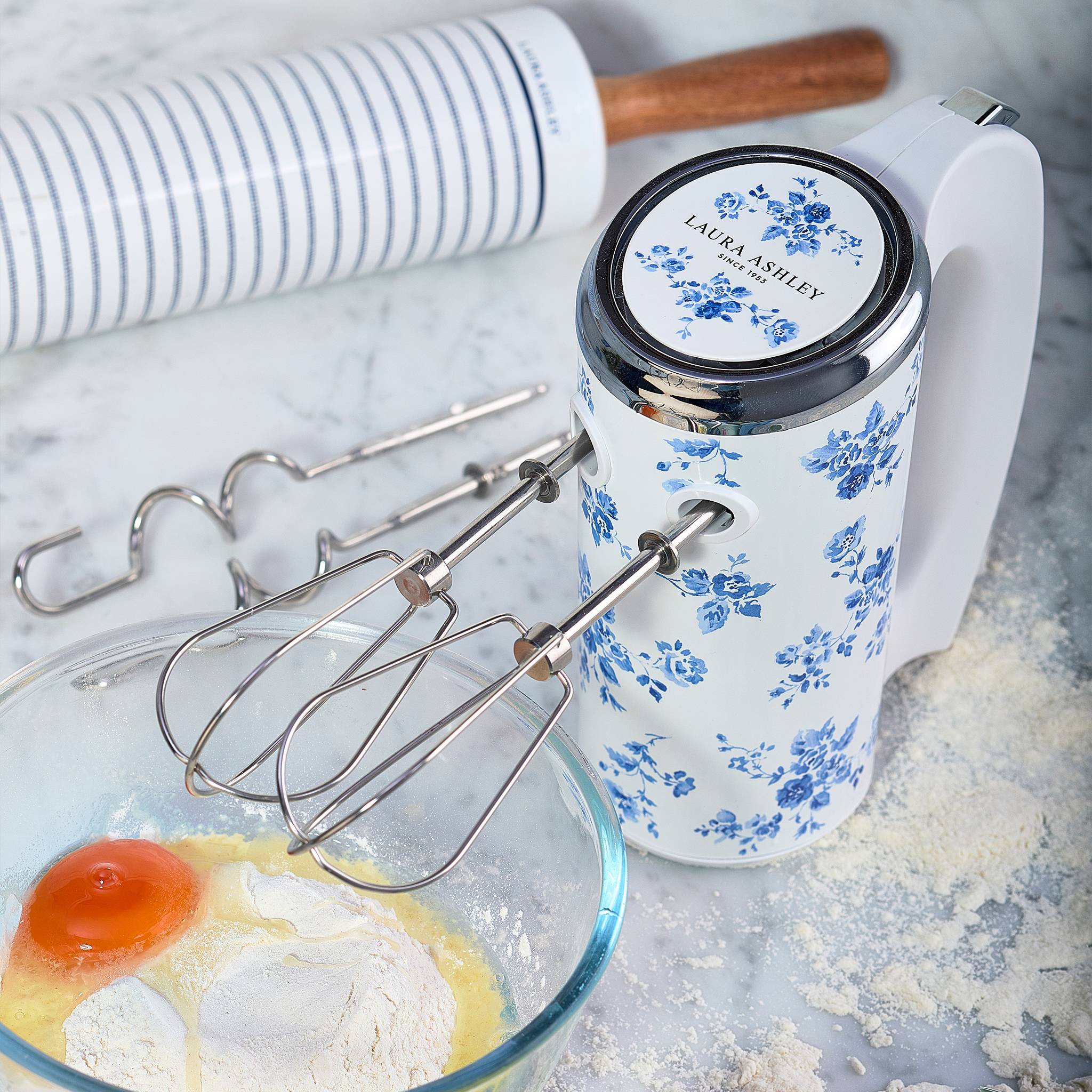 Ein weißer und blauer Handmixer mit Blumenmuster liegt neben einem Nudelholz auf einer mehligem Arbeitsplatte neben einer Schüssel mit Mehl, Butter und einem Ei.