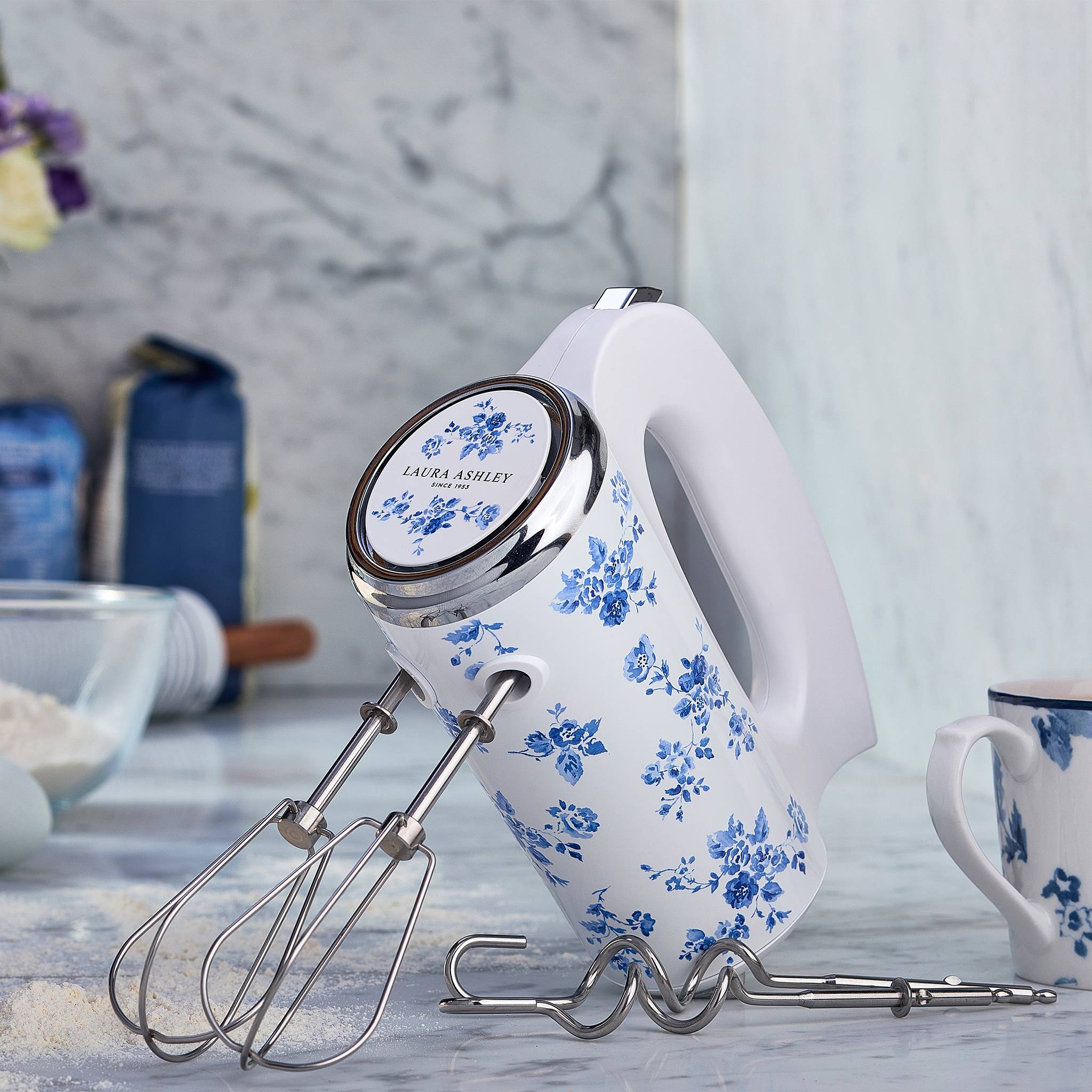 Ein weißer elektrischer Handmixer mit blauen Blütenmustern steht auf einer Küchentheke, umgeben von Backzutaten und Utensilien.