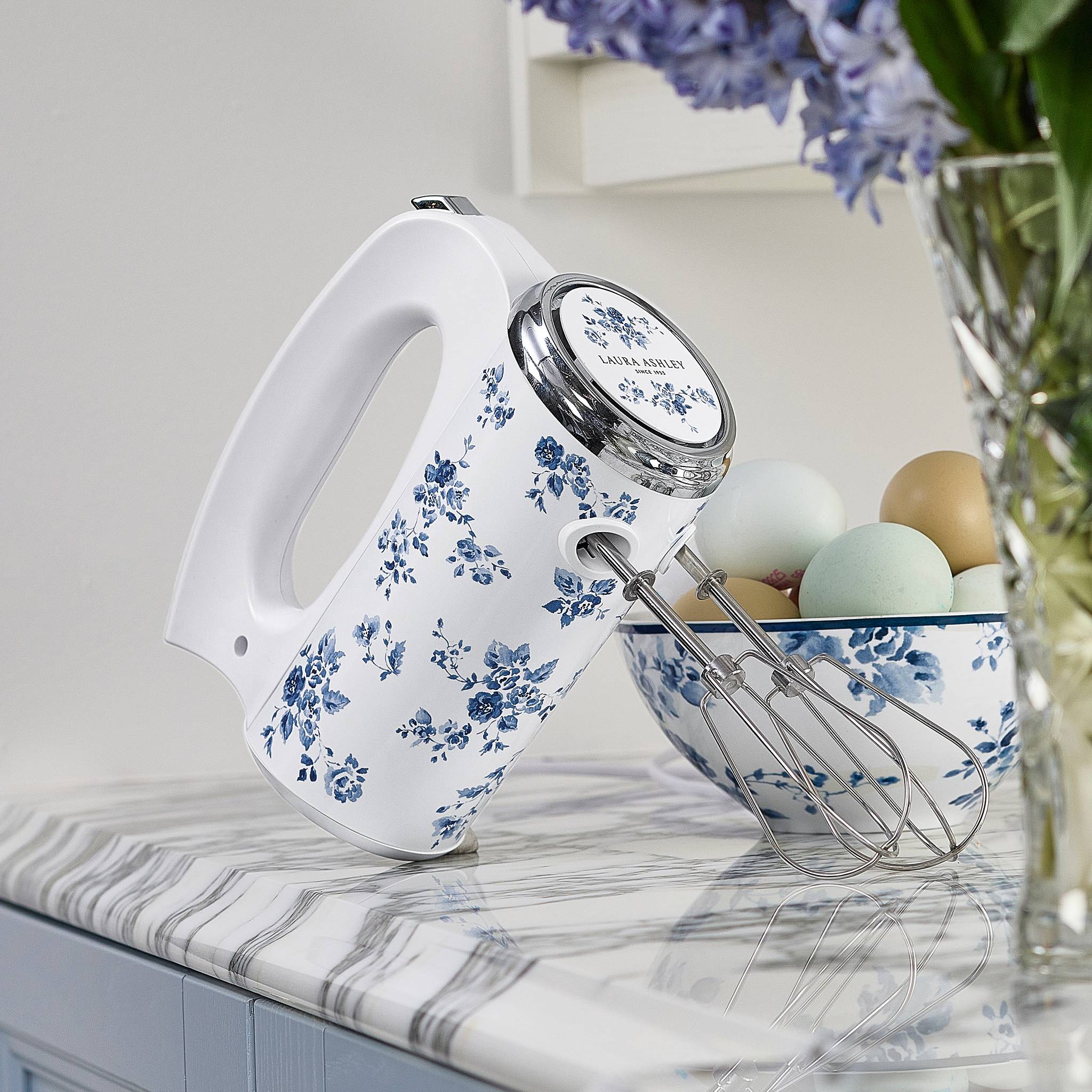 Ein elektrischer Mixer mit Blumenmuster ruht auf einer Marmorarbeitsplatte neben einer Schüssel mit Eiern, mit weichem Licht und einer gemütlichen Küchenumgebung.