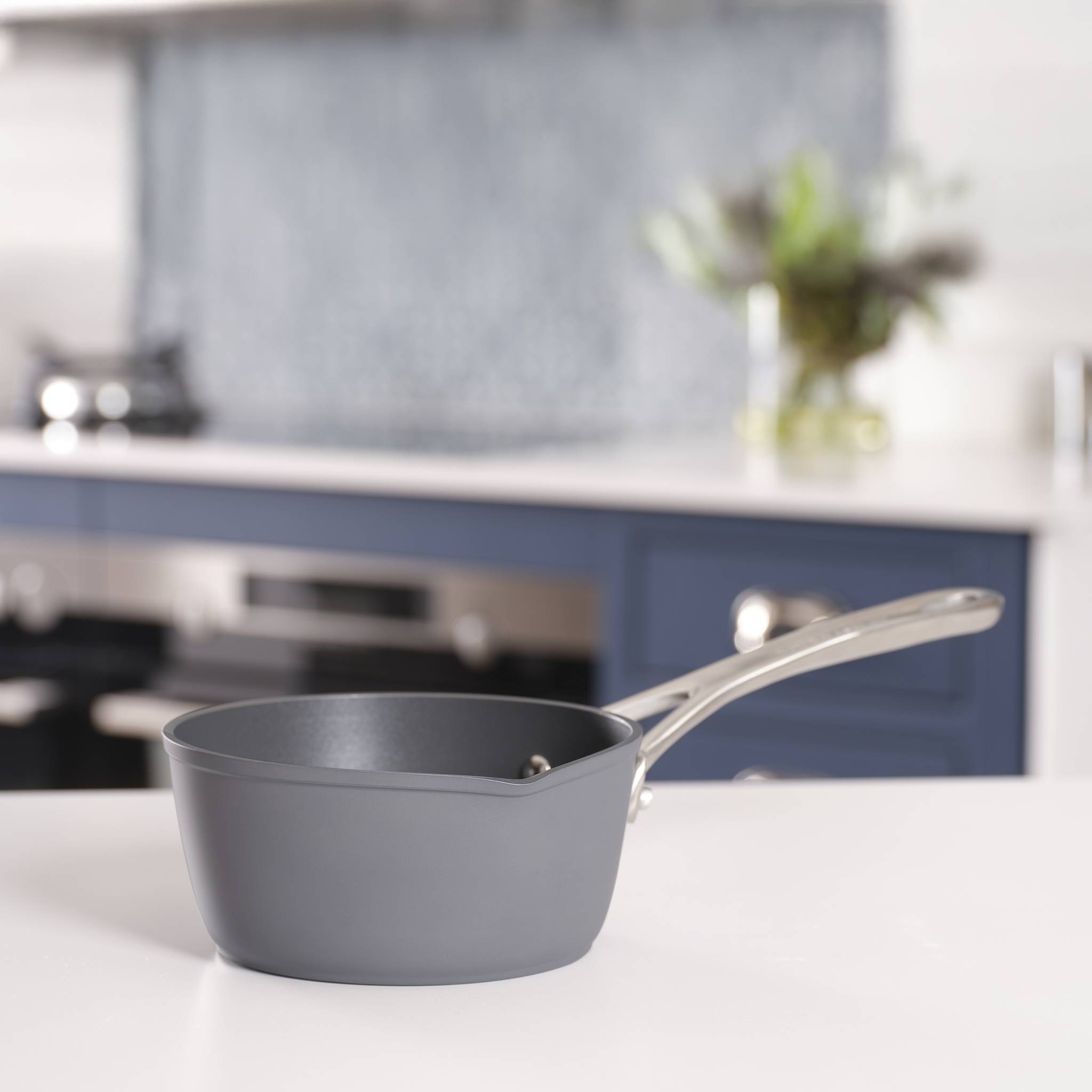 Eine graue Pfanne mit glänzendem Griff sitzt auf einer weißen Arbeitsplatte in einer modernen Kücheneinrichtung, unscharfer Hintergrund mit Pflanzen.