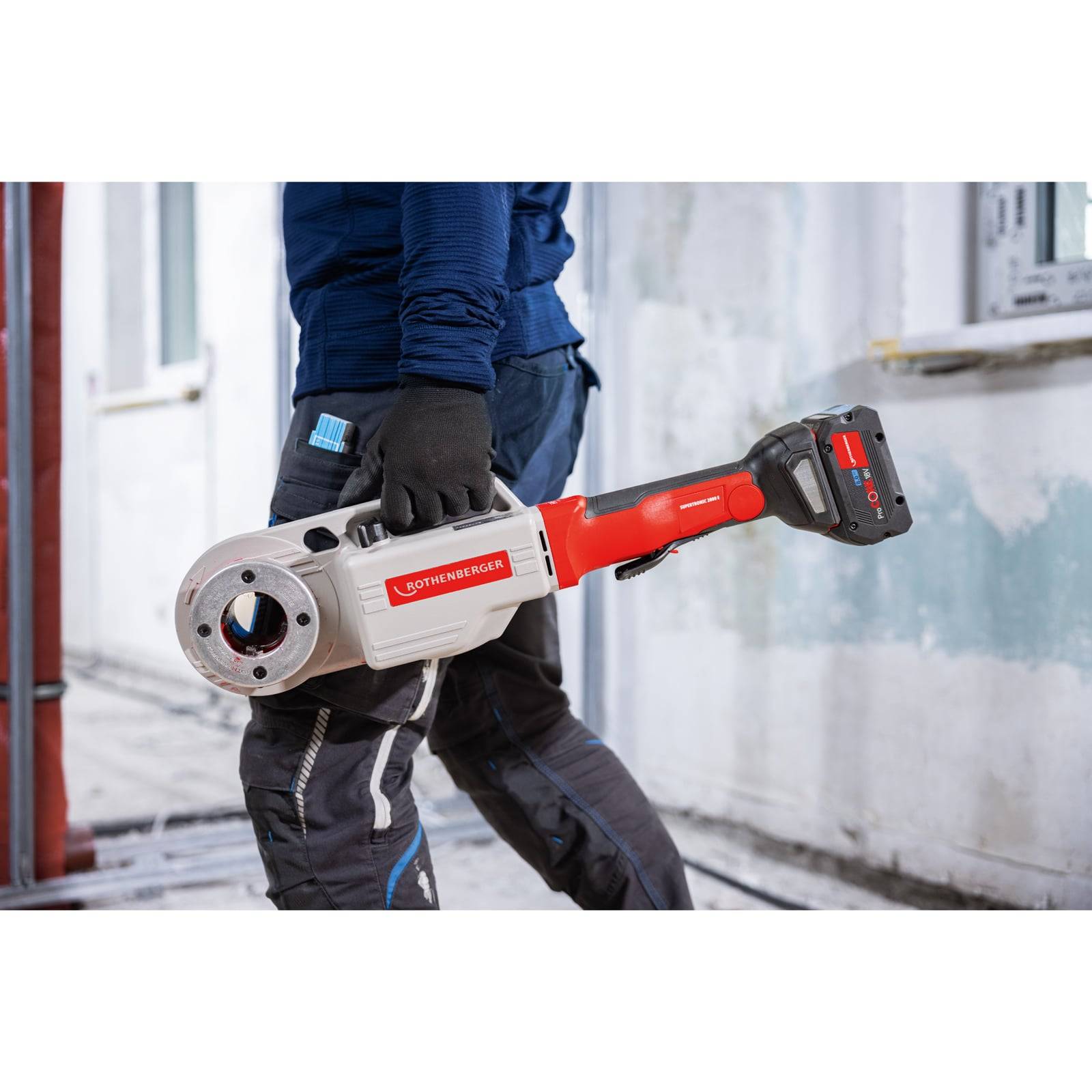 Eine Person, die ein rotes und silbernes tragbares Rohrzeugsortiment in Innenräumen verwendet, trägt Schutzhandschuhe und blaue Arbeitskleidung in einer Baustelle.