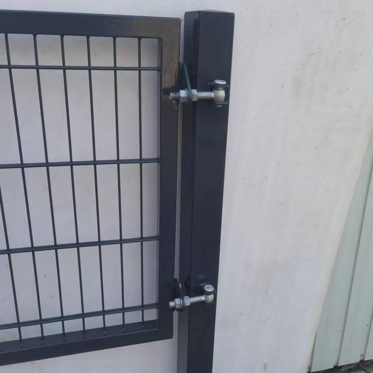 Ein Metalltor mit vertikalen und horizontalen Stäben ist mit Scharnieren an einem stabilen Pfosten befestigt. Die Konstruktion befindet sich vor einer hellfarbigen Wand.