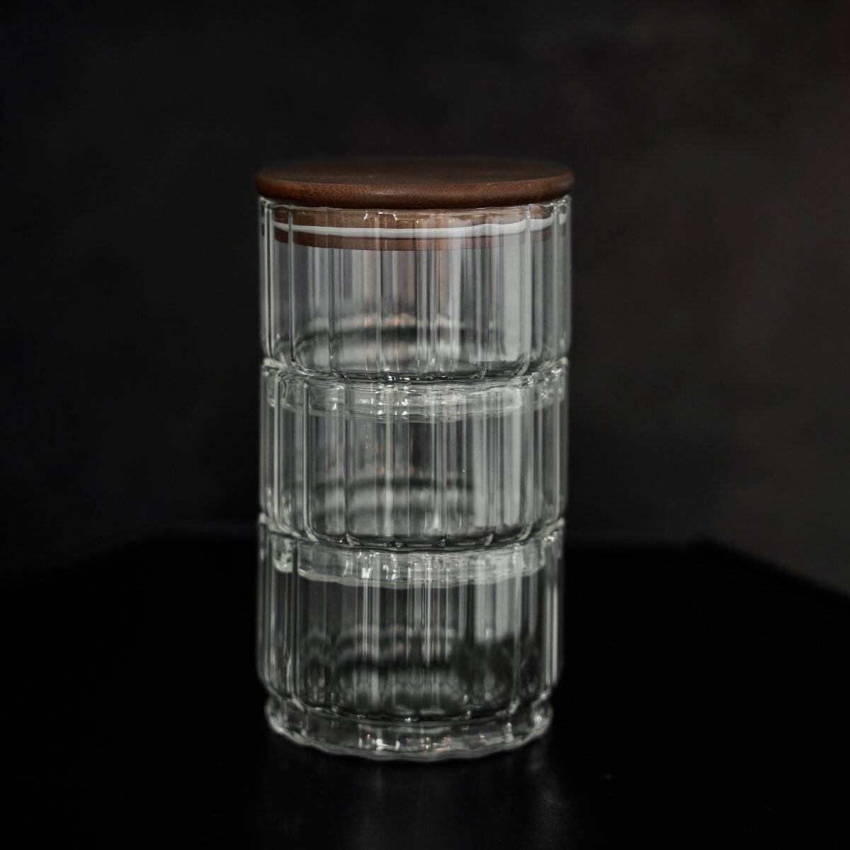 Ein Glasbehälter mit vertikalen Nuten und einem Holzdeckel vor dunklem Hintergrund.