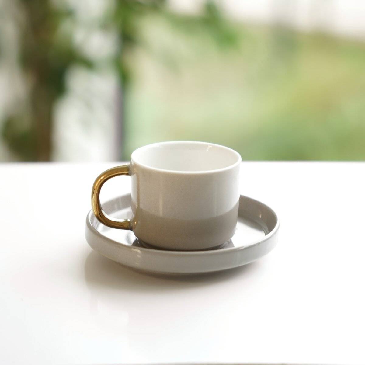 Eine weiße Kaffeetasse mit goldenem Griff sitzt auf einer passenden Untertasse auf einem weißen Tisch, mit einem unscharfen grünen Hintergrund dahinter.