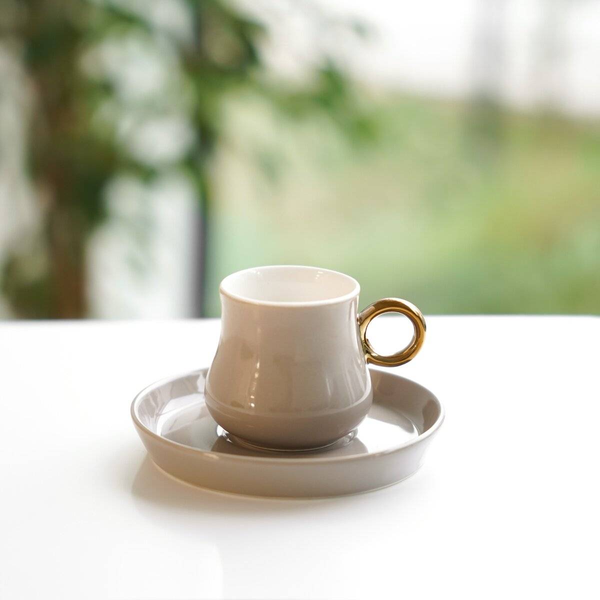 Eine weiße Keramiktasse mit einem einzigartigen abgerundeten Henkel sitzt auf einem asymmetrischen Tablett vor einem schlichten weißen Hintergrund.