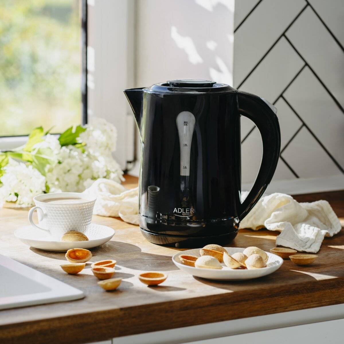 Ein schwarzer Wasserkocher auf einer Küchentheke neben einer Tasse Tee, Keksen und einem weißen Tuch, mit einem Fenster und Blumen im Hintergrund.