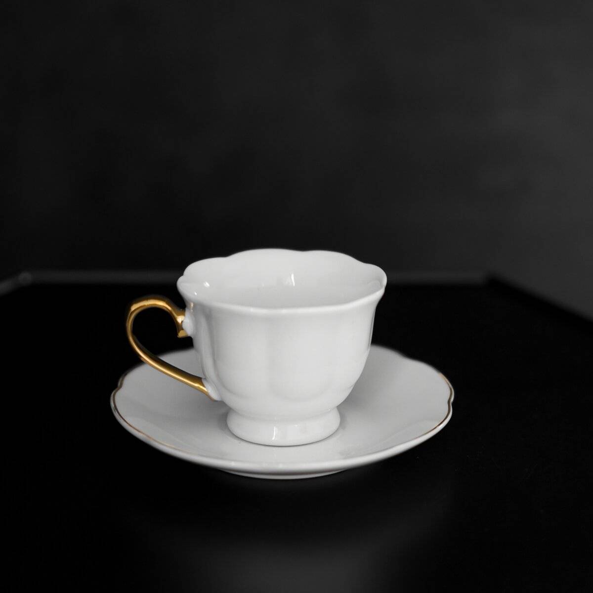 Eine weiße Teetasse mit goldenem Griff sitzt auf einer passenden Untertasse vor dunklem Hintergrund.