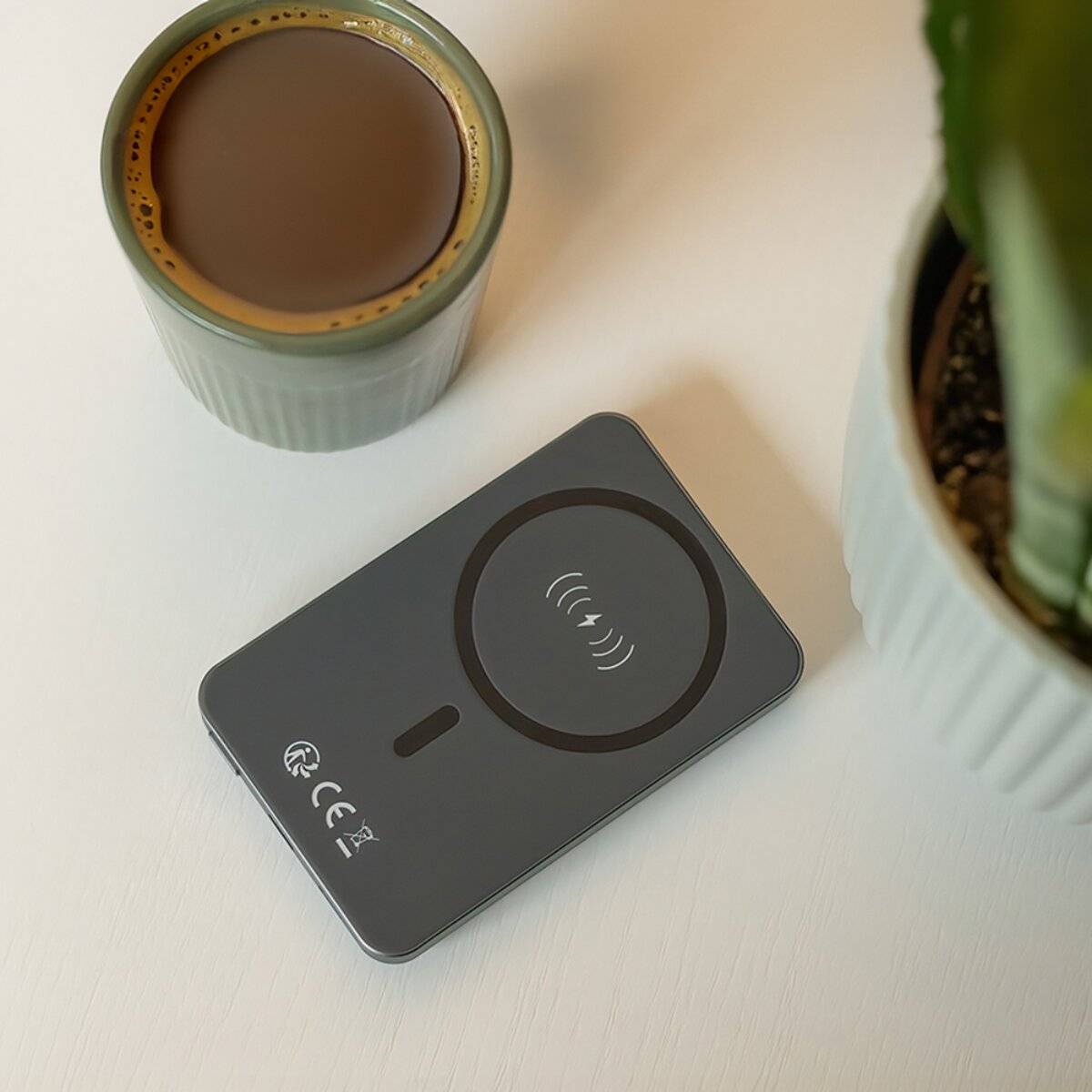 Ein kabelloses Ladegerät auf einem Tisch neben einer vollen Kaffeetasse und einer Pflanze, das moderne technische Bequemlichkeit im Alltag veranschaulicht.