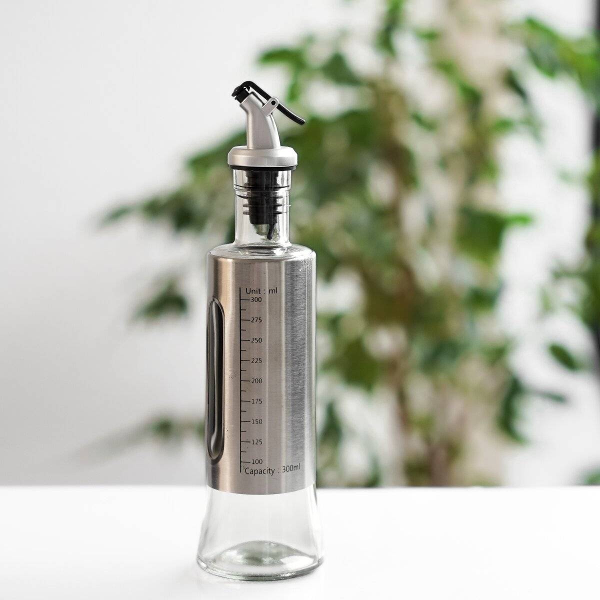 Eine Glasölflasche mit Spender-Aufsatz aus Edelstahl und Messskalen auf einem Tisch. Eine unscharfe Pflanze ist im Hintergrund zu sehen.