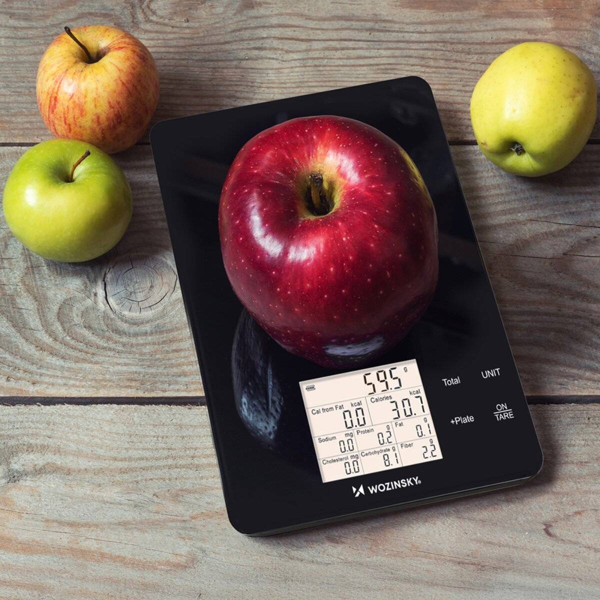 Ein roter Apfel wiegt 595 Gramm auf einer digitalen Küchenwaage, umgeben von zwei grünen Äpfeln und einem gelben Apfel auf einer Holzoberfläche.