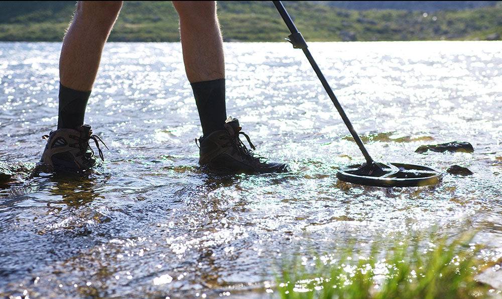 Eine Person in Stiefeln benutzt ein Metalldetektor, um in flachem Wasser an einem sonnigen Tag nach Objekten zu suchen.