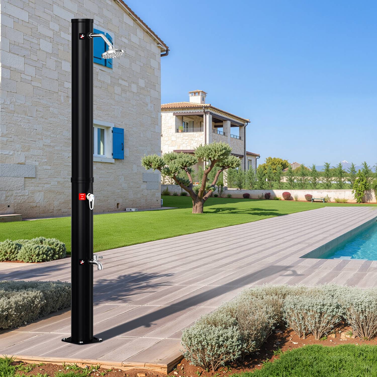 Eine schwarze Außendusche steht auf einer gefliesten Terrasse mit einem üppig grünen Rasen und einem großen Steinhaus im Hintergrund unter einem klaren Himmel.