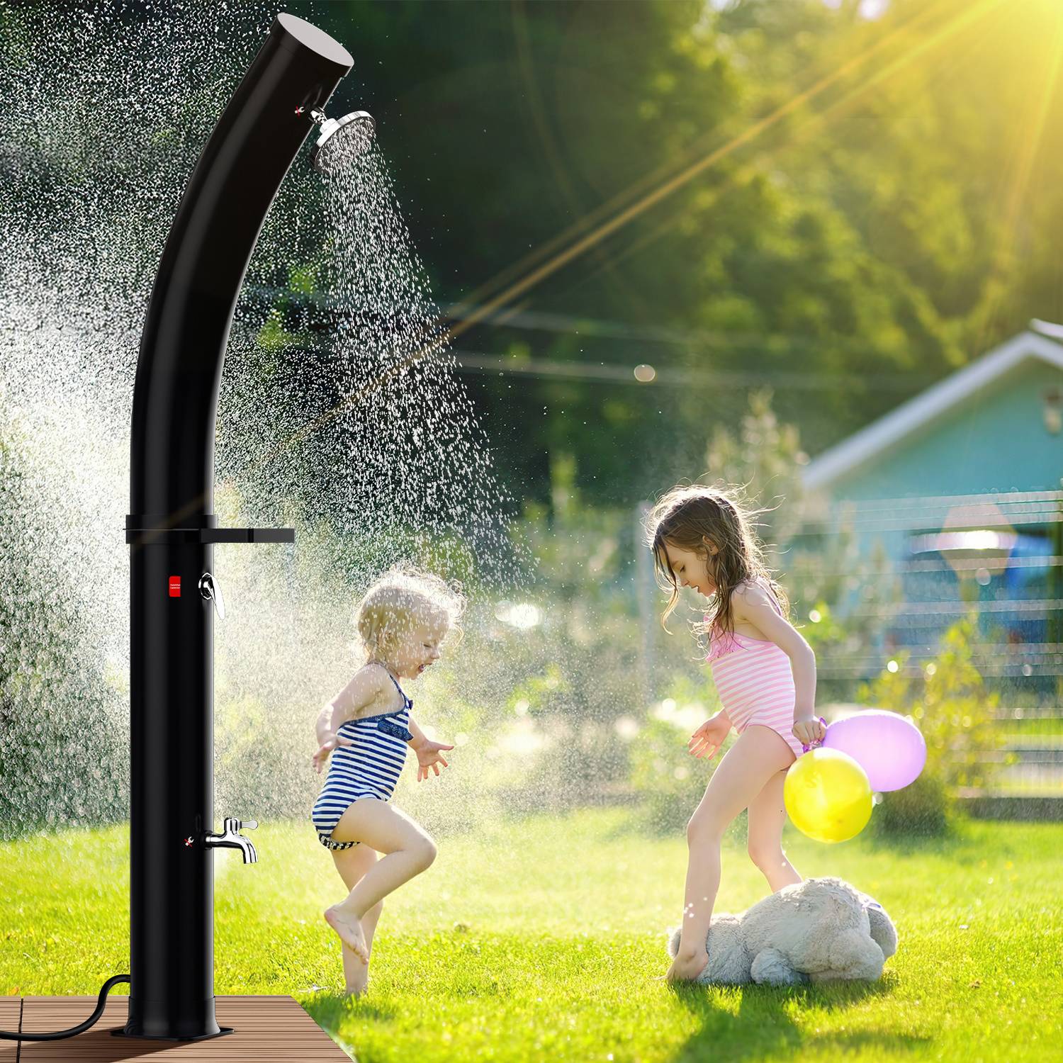 Zwei Kinder spielen fröhlich unter einer Außendusche an einem sonnigen Tag, wobei ein Kind Luftballons hält und grünes Gras im Hintergrund ist.
