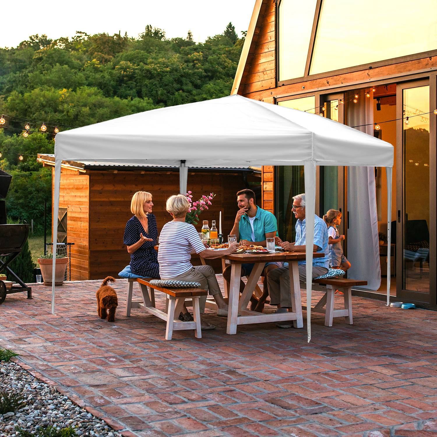 Eine Gruppe von Menschen sitzt um einen Außenesstisch unter einem Baldachin und genießt eine Mahlzeit auf einer Terrasse mit Wald und Haus im Hintergrund.