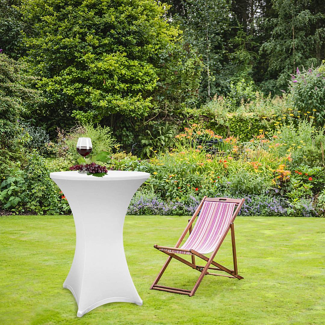 Ein Garten mit einem hohen Tisch, der mit weißem Tuch drapiert ist und ein Glas Rotwein hält, neben einem gestreiften Liegestuhl auf grünem Gras.