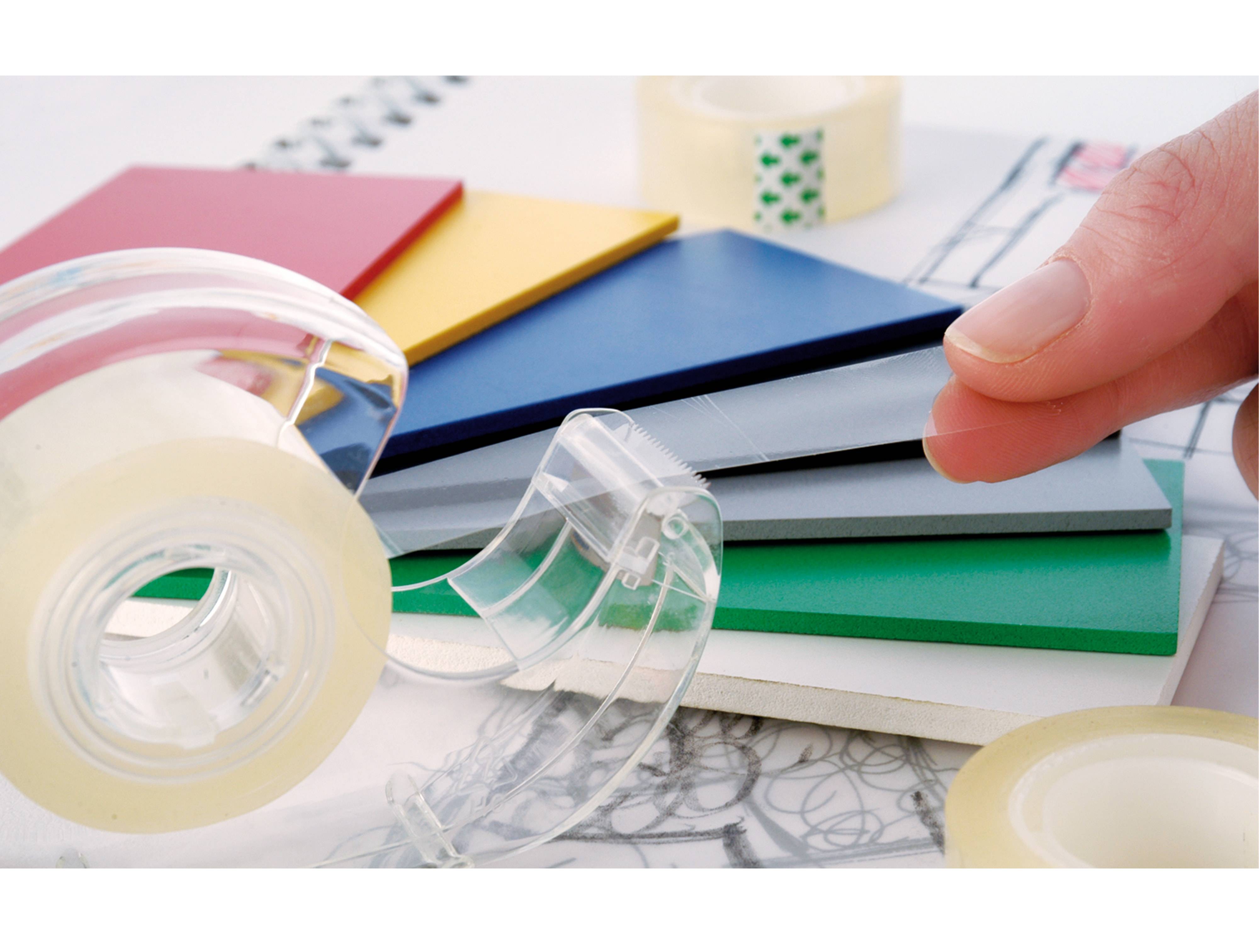 Eine Hand, die Klebeband von einem Spender neben bunten gestapelten Ordnern und einem Notizbuch abzieht, was auf eine Organisations- oder Büroumgebung hindeutet.