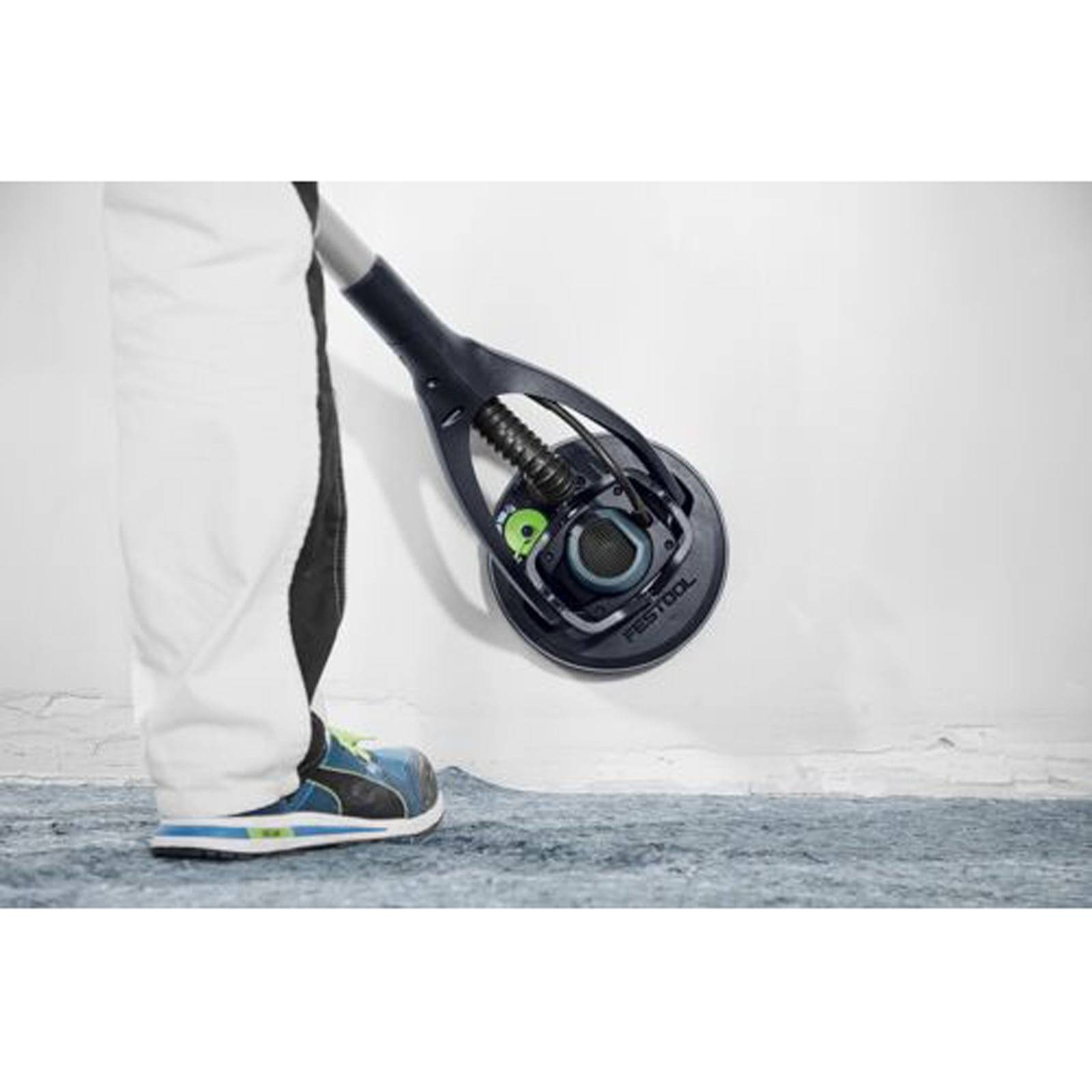 Eine Person benutzt einen Elektroschleiffer an einer weißen Wand. Die Person trägt weiße Hosen mit einem schwarzen Streifen und schwarze Sneaker mit blauen und gelben Akzenten.