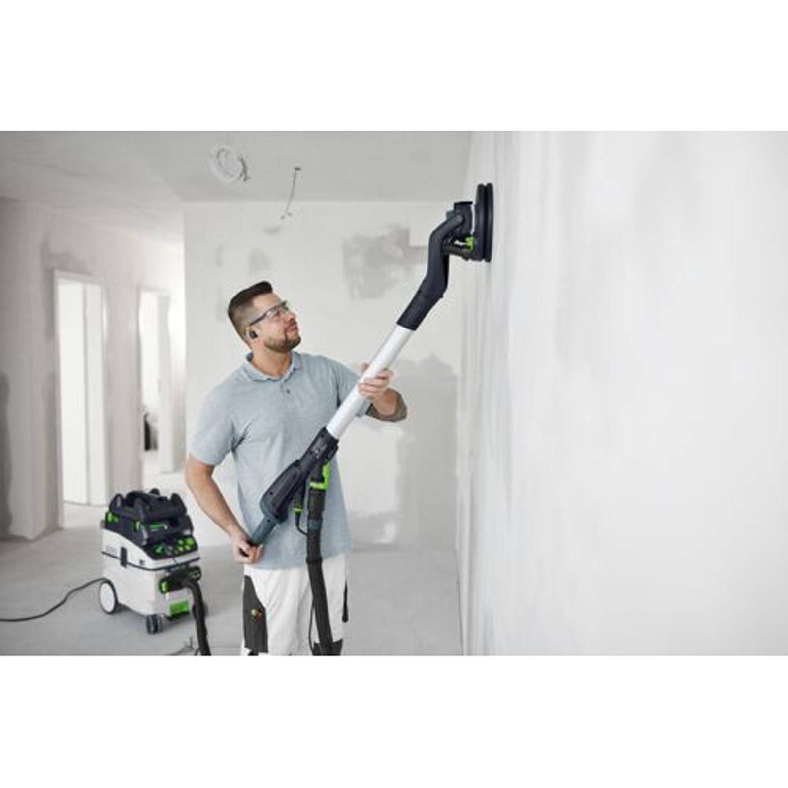 Eine Person nutzt einen Elektroschleiffer, um eine Wand in einem Raum unter Renovierung zu glätten, wobei ein Staubsauger mit dem Schleiffer für die Staubsammlung verbunden ist.