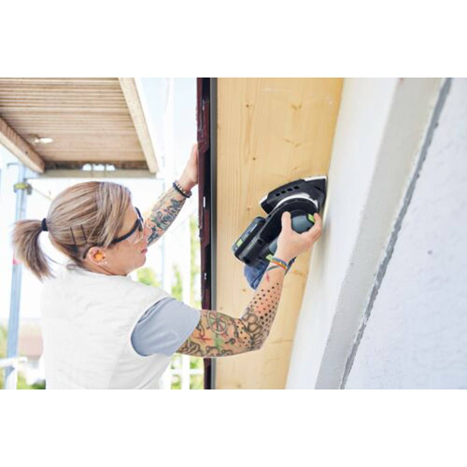 Eine Person mit Tätowierungen und Sonnenbrille benutzt ein Elektrowerkzeug an holzerner Verkleidung, während sie auf einem Gerüst neben einem teilweise fertiggestellten Gebäude steht.