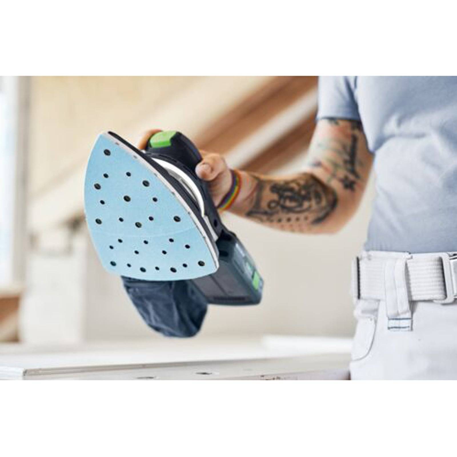 Person, die einen tragbaren Elektroschlifer hält, mit sichtbaren Tätowierungen am Arm, bereitet sich darauf vor, eine Oberfläche in einer Inneneinstellung abzuschleifen.