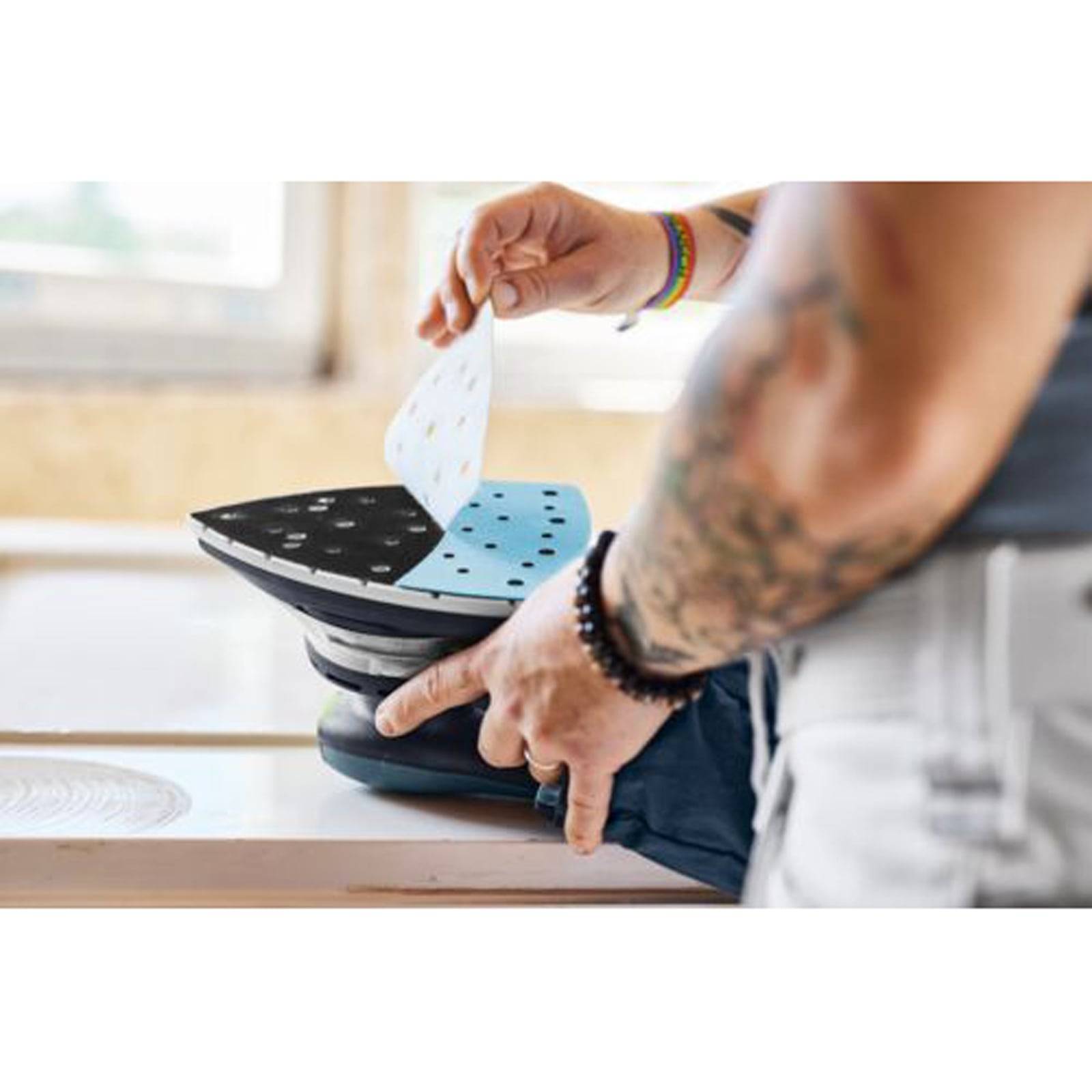 Eine Person wechselt Schleifpapier an einem elektrischen Schleiffer in einer Werkstatt. Ihr Unterarm ist mit Tätowierungen und Armbändern sichtbar.