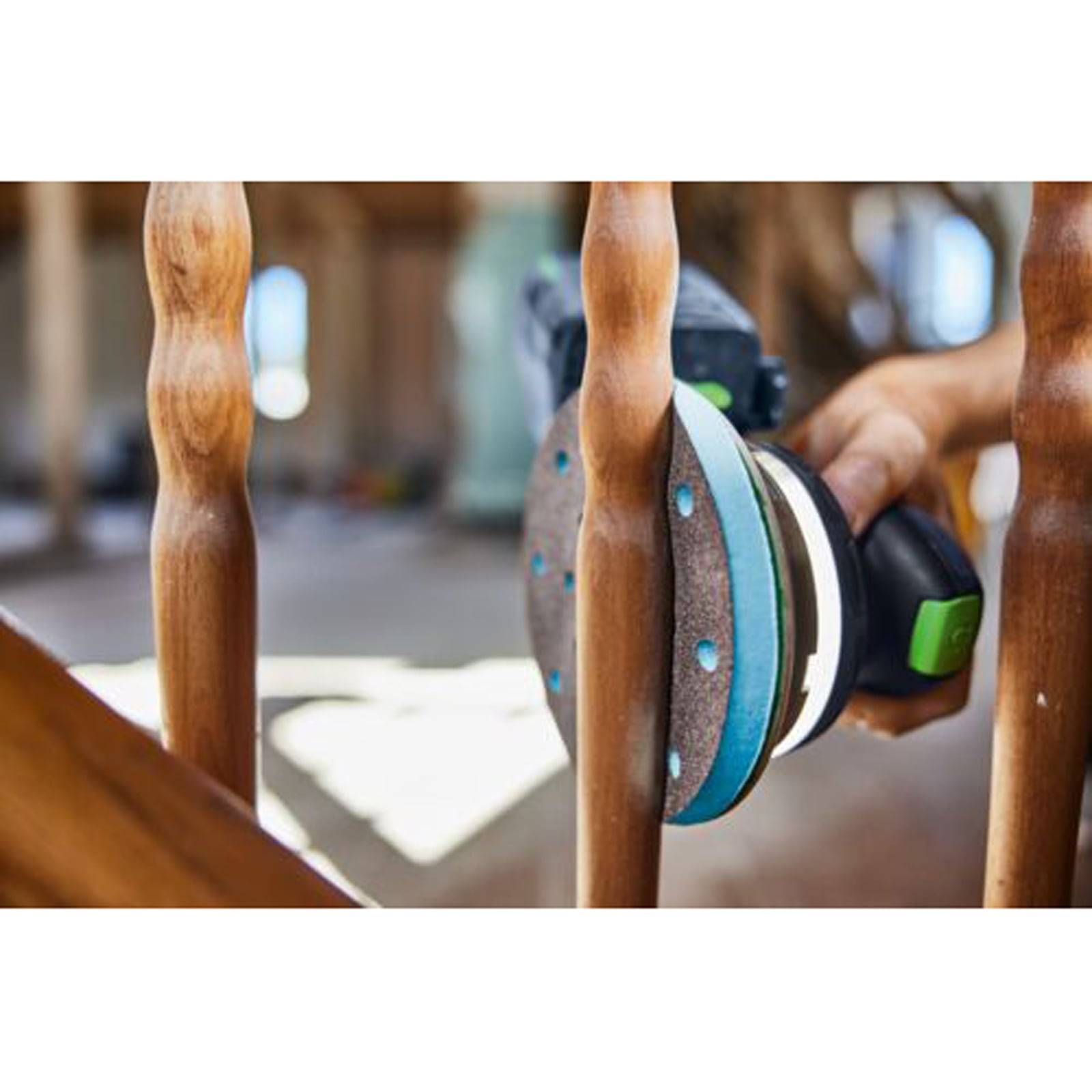 Eine Person benutzt einen elektrischen Schleiffer auf einem Holzstuhl und glättet die Oberfläche. Der Hintergrund zeigt einen unscharfen Innenraum.