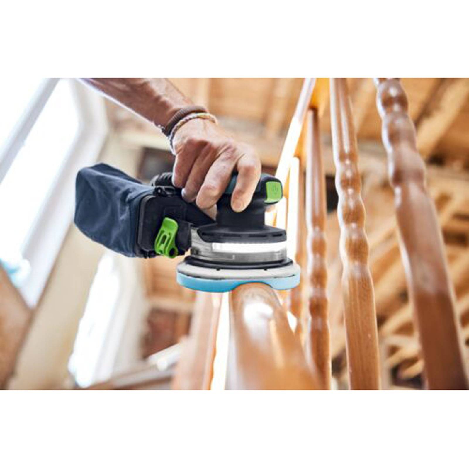 Eine Person verwendet einen tragbaren elektrischen Schleiffer, um ein hölzernes Geländer in Innenräumen zu glätten, wobei eine Renovierungs- oder Holzbearbeitungsaktivität im Haushalt hervorgehoben wird.