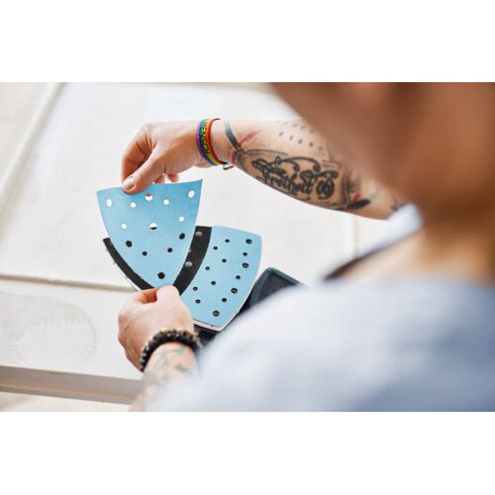 Eine Person mit tätowierten Armen hält zwei durchlöcherte, dreieckige Schleifblätter und bereitet sich darauf vor, diese an einem Schleiflipwerkzeug zu befestigen, in einer Werkstattumgebung.