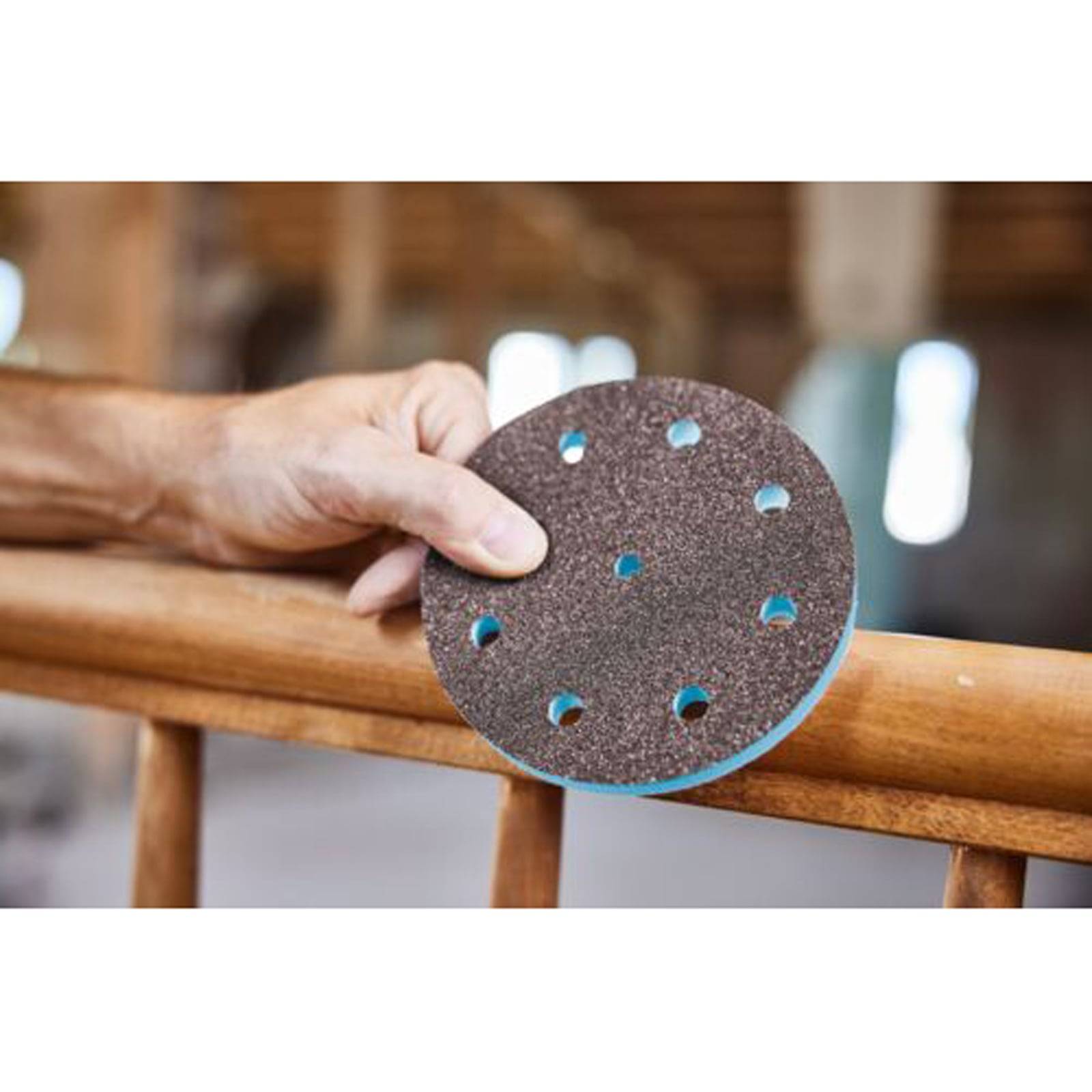 Eine Hand hält eine kreisförmige Schleifscheibe mit Löchern, die über einer Holzoberfläche positioniert ist, was auf die Vorbereitung zum Schleifen oder Aufarbeiten hindeutet.