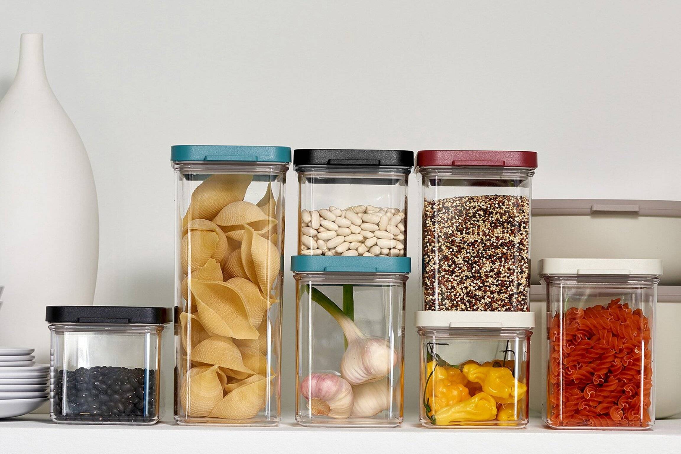 Verschiedene Trockenfood ordentlich in fünf durchsichtigen Behältern auf einem Regal organisiert, darunter Nudeln, Bohnen, Quinoa, Knoblauch und Paprika.