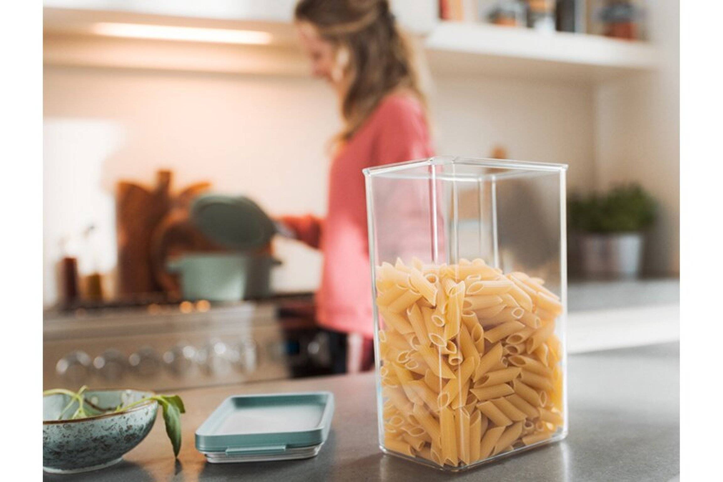 Ein klarer Behälter gefüllt mit Penne-Pasta steht auf einer Küchentheke; eine Person im Hintergrund, leicht unscharf, kocht.