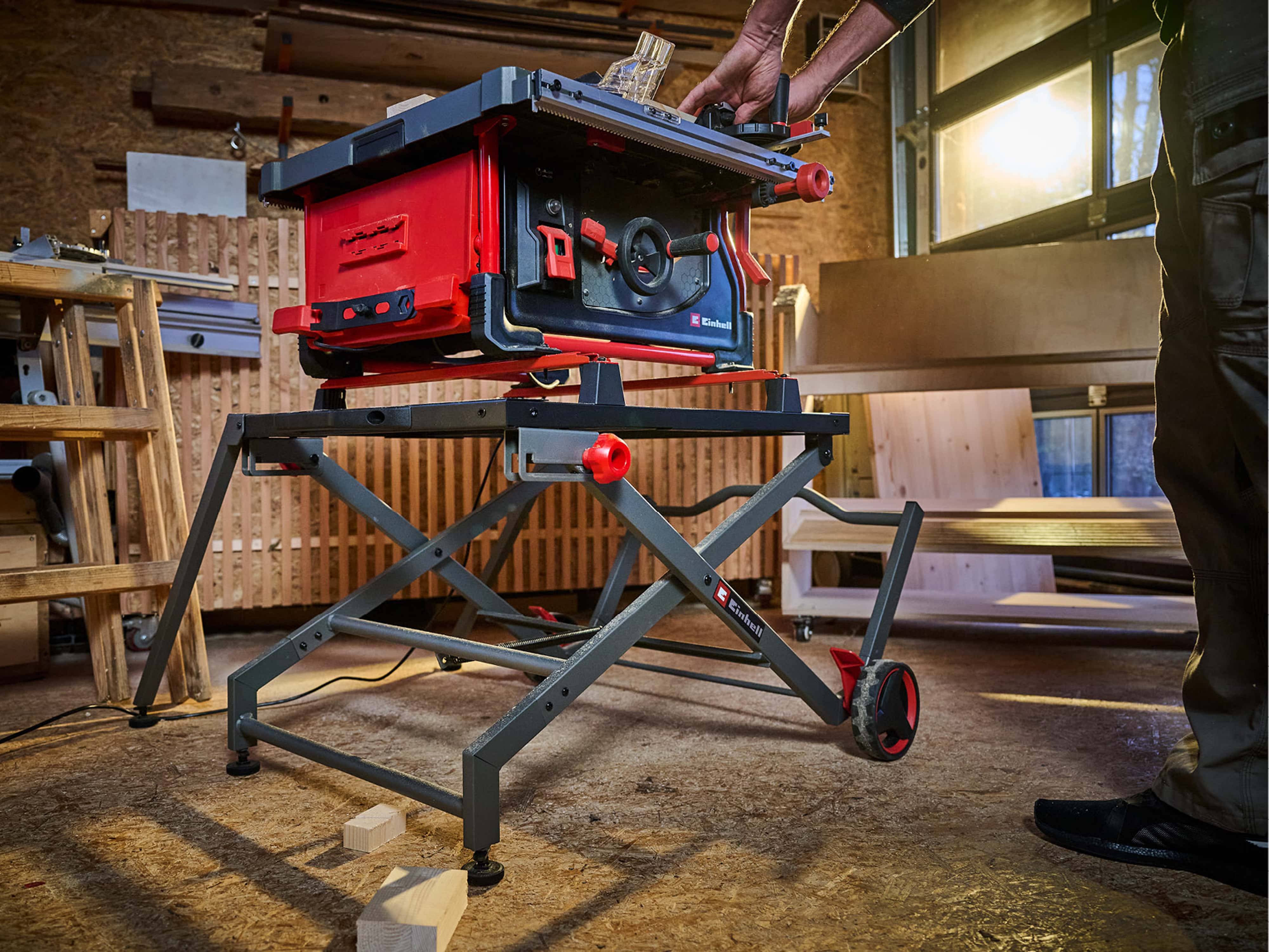 Eine Person stellt eine rot-schwarze Tischkreissäge auf einem Metallständer in einer Werkstatt mit Holzmaterialien und Werkzeugen herum ein.