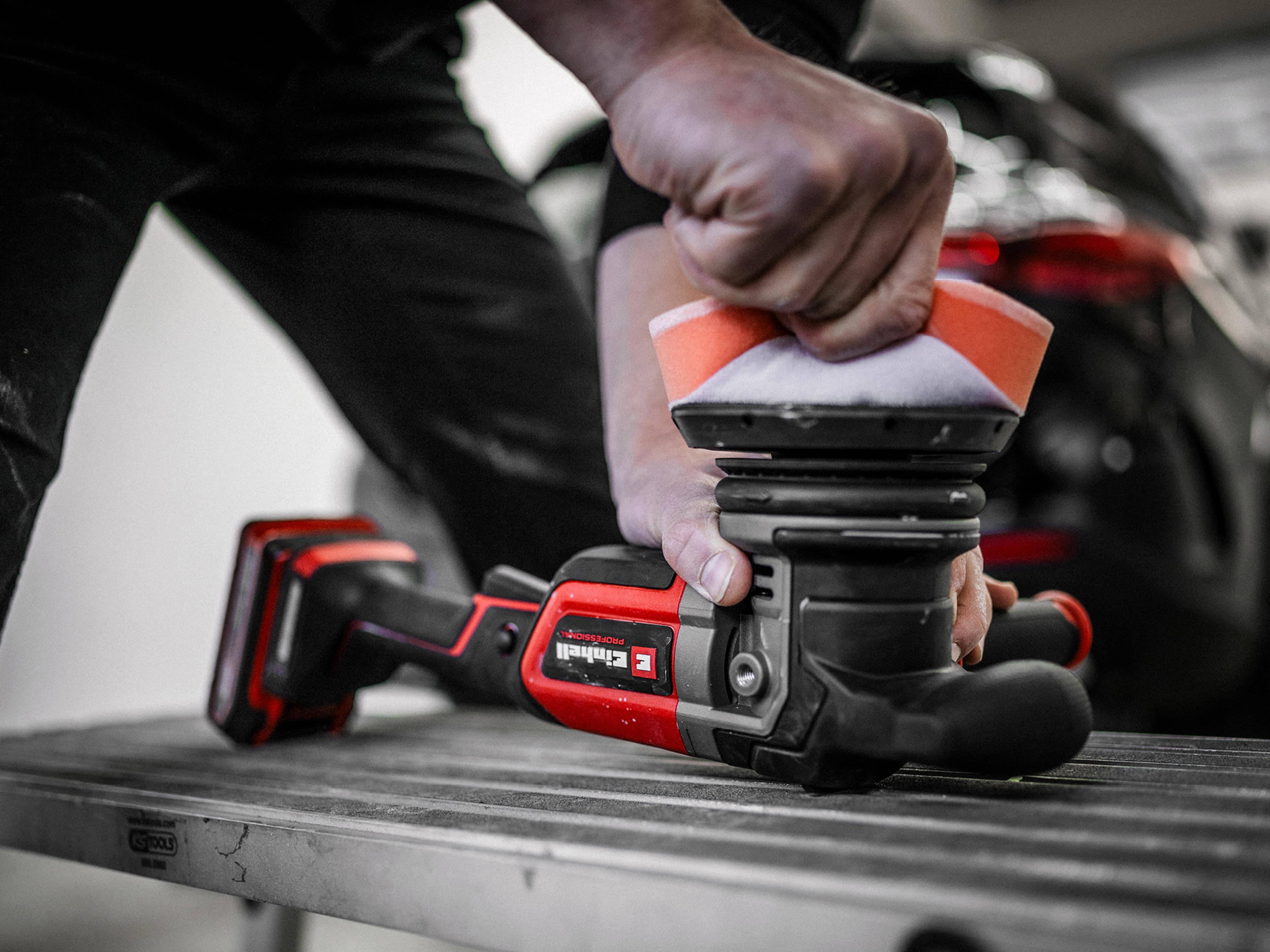 Eine Person, die einen rot-schwarzen elektrischen Schleiffer auf einer Holzoberfläche verwendet, mit Fokus auf ihre Hand, die das Werkzeug greift, wobei seine Verwendung hervorgehoben wird.