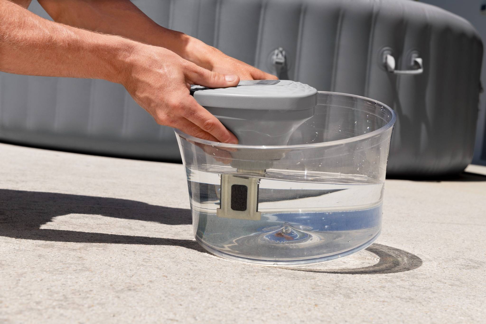 Eine Hand hält ein tragbares Gerät in einer klaren Schüssel, die mit Wasser gefüllt ist, auf einer Betonoberfläche positioniert, mit einem aufblasbaren Pool im Hintergrund.