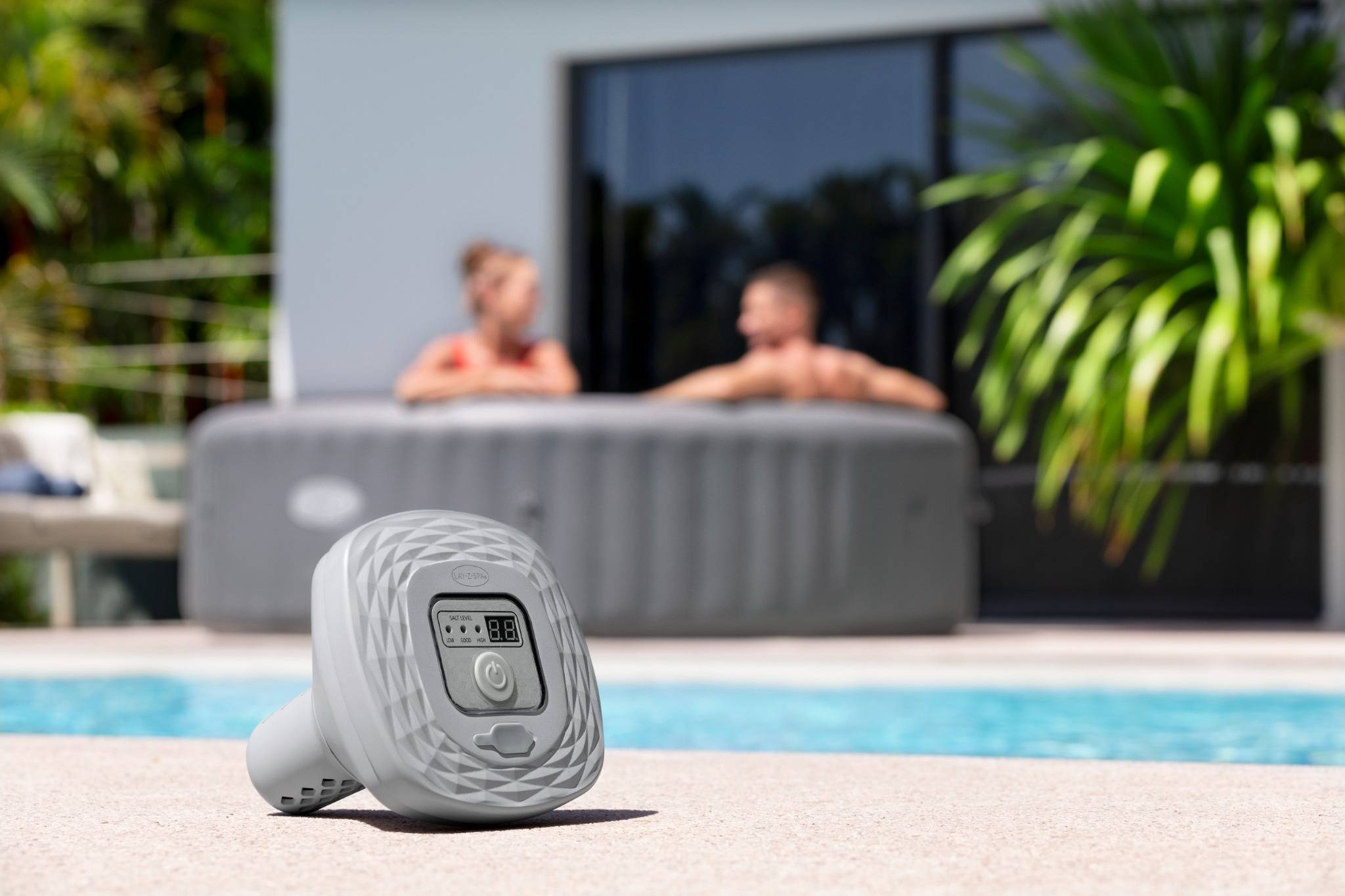 Ein digitales Poolthermometer am Poolrand mit zwei unscharfen Personen in einem Whirlpool im Hintergrund, was eine entspannende Außenumgebung andeutet.