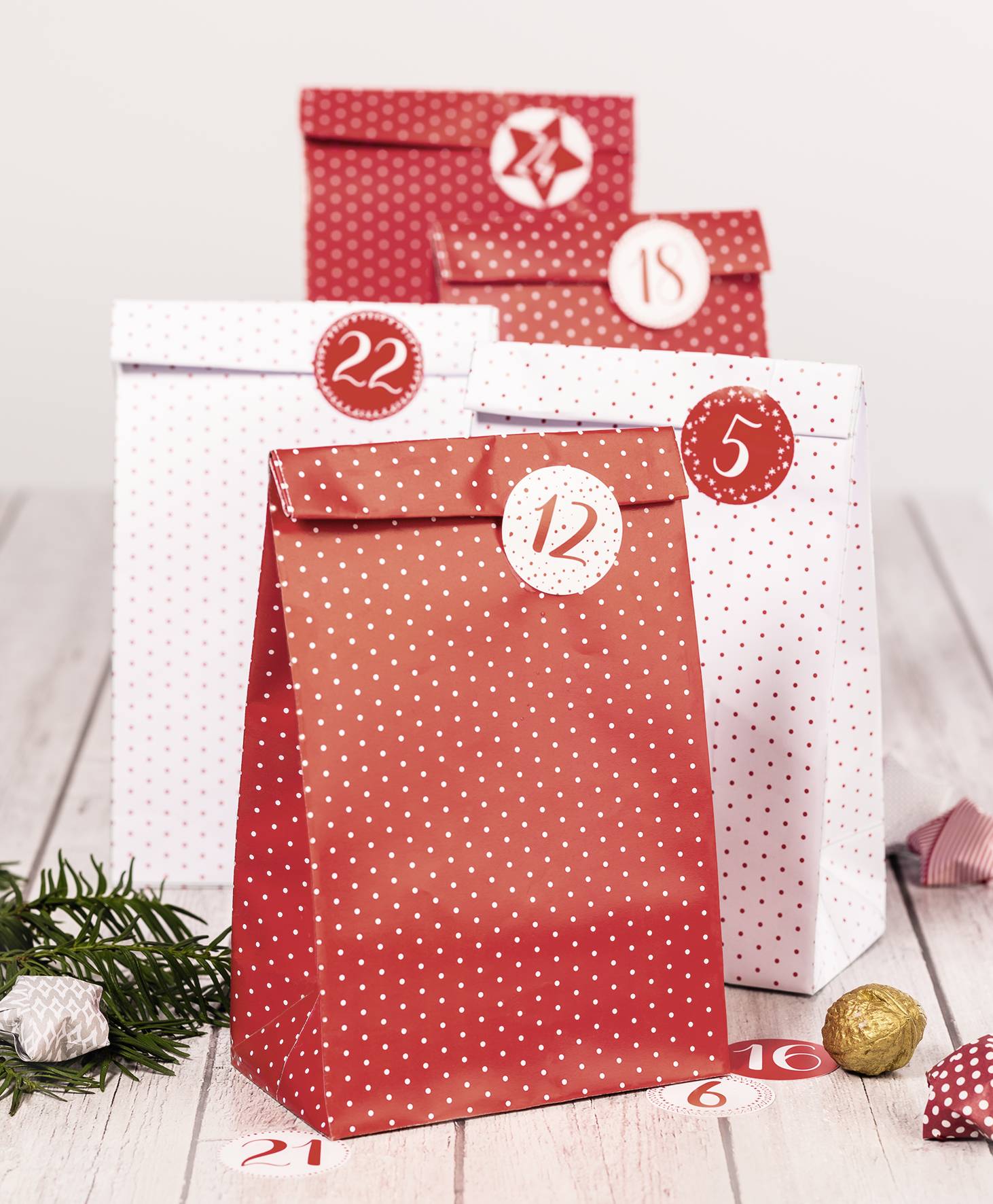 Weihnachtliche Adventskalender-Szene mit rot-weißen gepunkteten Tüten mit den Nummern 5, 12, 15, 22, umgeben von Kiefernzweigen und Weihnachtskugeln.