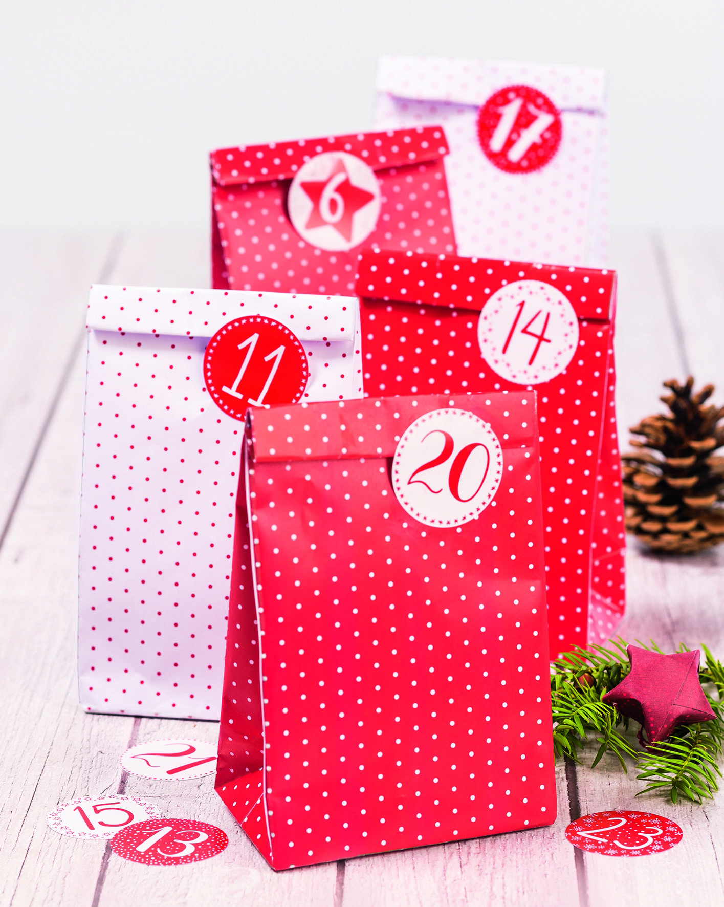 Satz aus roten und weißen Weihnachtsgeschenktüten mit den Nummern 6, 11, 14 und 20, verziert mit Punkten, Tannenzapfen und Tannenzweigen auf einer Holzoberfläche.