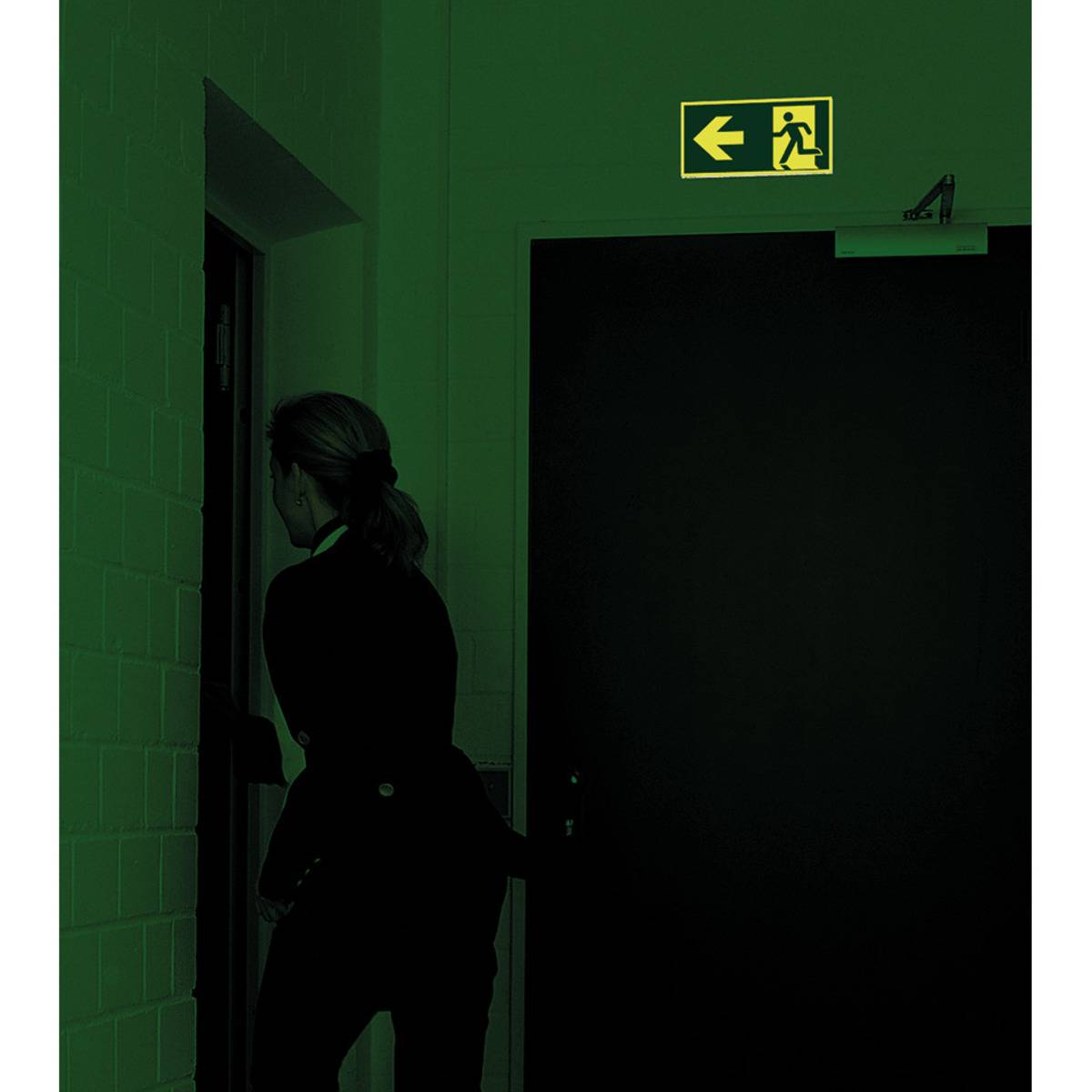Eine Person öffnet eine Tür unter einem beleuchteten Notausgangsschild, das einen Pfeil und eine laufende Figur zeigt und eine Notausgansroute anzeigt.
