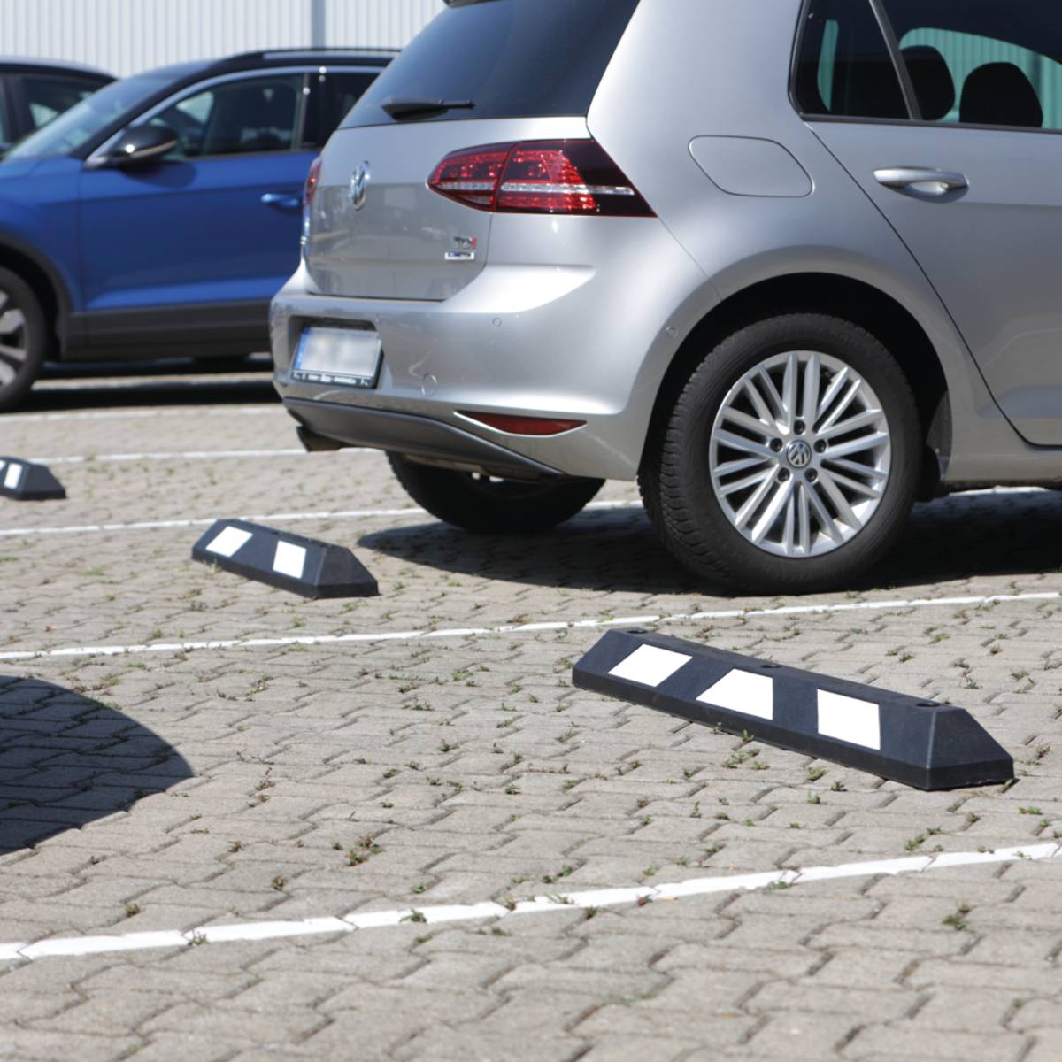 Ein silberner Wagen parkt in einem gekennzeichneten Platz mit Radstoppern; ein blauer Wagen ist im Hintergrund.