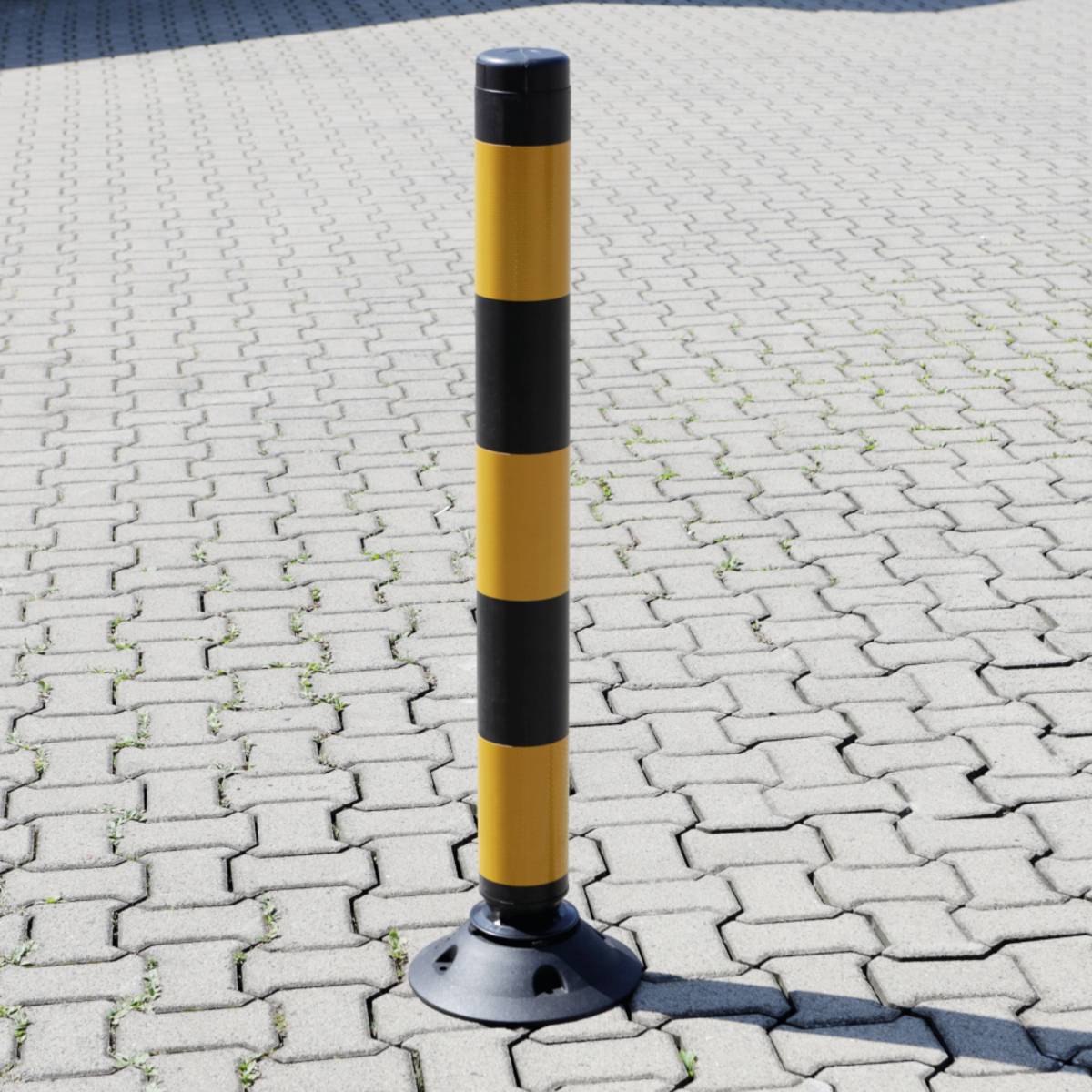 Ein schwarz-gelb gestreifter Poller steht auf einem sonnigen Backsteinbelag.