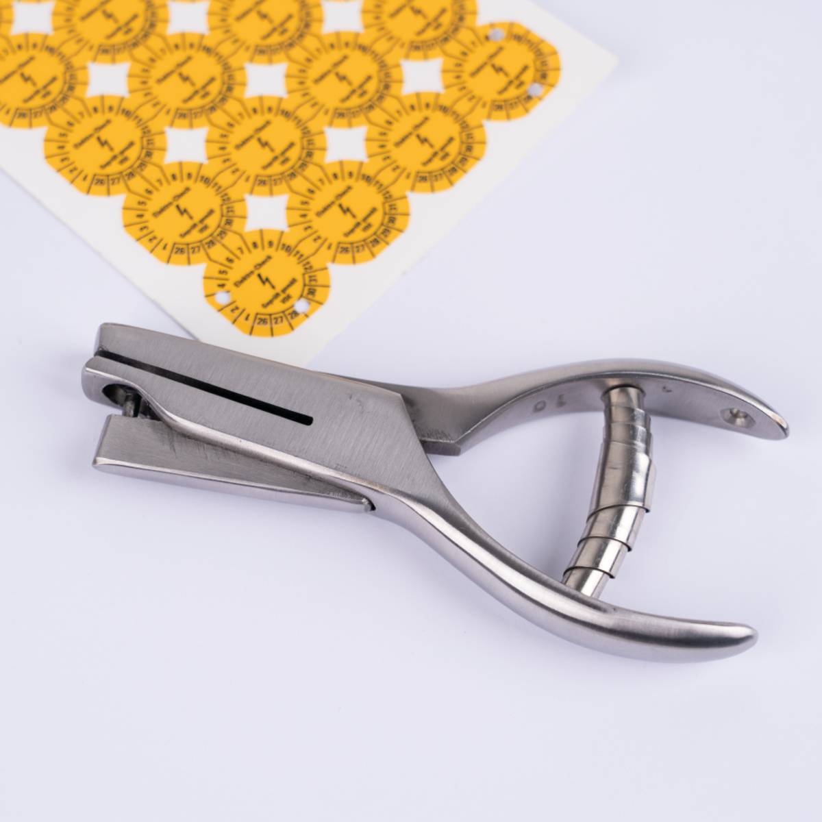 Stahlösenlocher-Werkzeug neben gelbem Papierblatt mit mehreren kreisförmigen Skalierungsmustern. Wird zum Einsetzen von Ösen in Materialien verwendet.