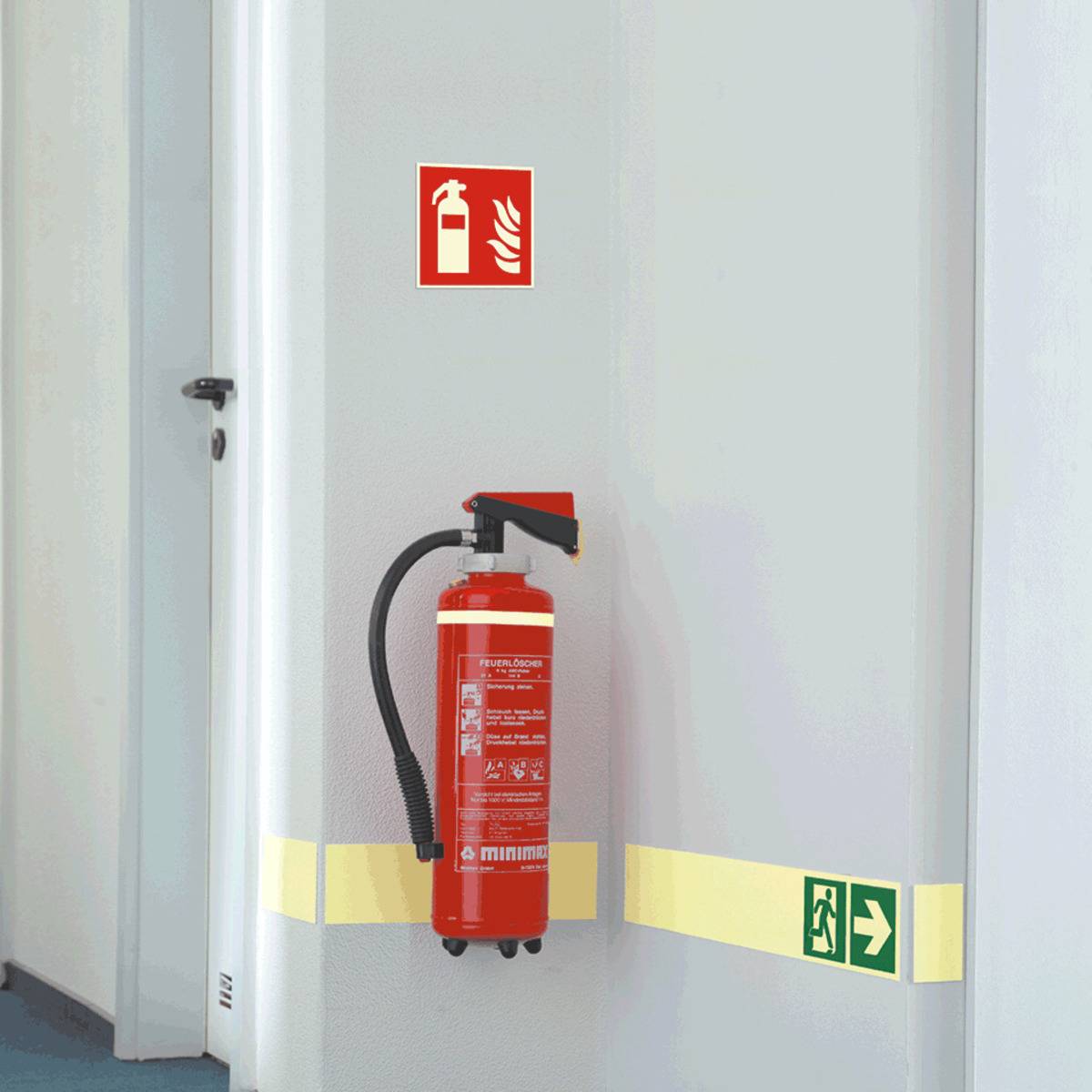Ein Feuerlöscher an der Wand unter einem Brandschutzschild angebracht, neben einer Tür in einem Flur, mit einem Notausgangsschild in der Nähe.