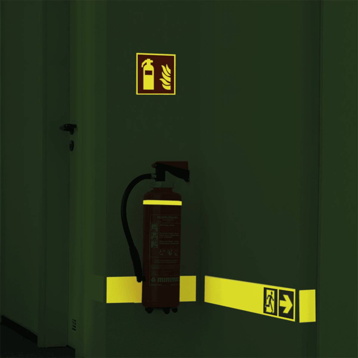 Ein Feuerlöscher ist an einer Wand angebracht, in der Nähe befinden sich selbstleuchtende Brandschutzzeichen, einschließlich eines Richtungsausgangsschildes.