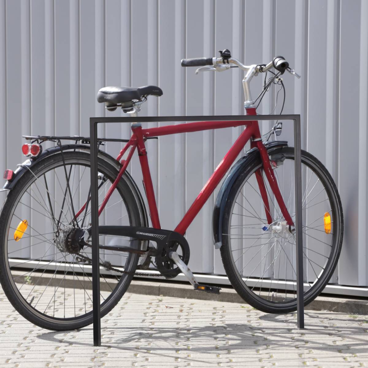 Ein rotes Fahrrad ist an einem Metallfahrradständer auf einer gepflasterten Oberfläche gegen eine wellblechverstärkte Metallwand in einer städtischen Umgebung gesichert.
