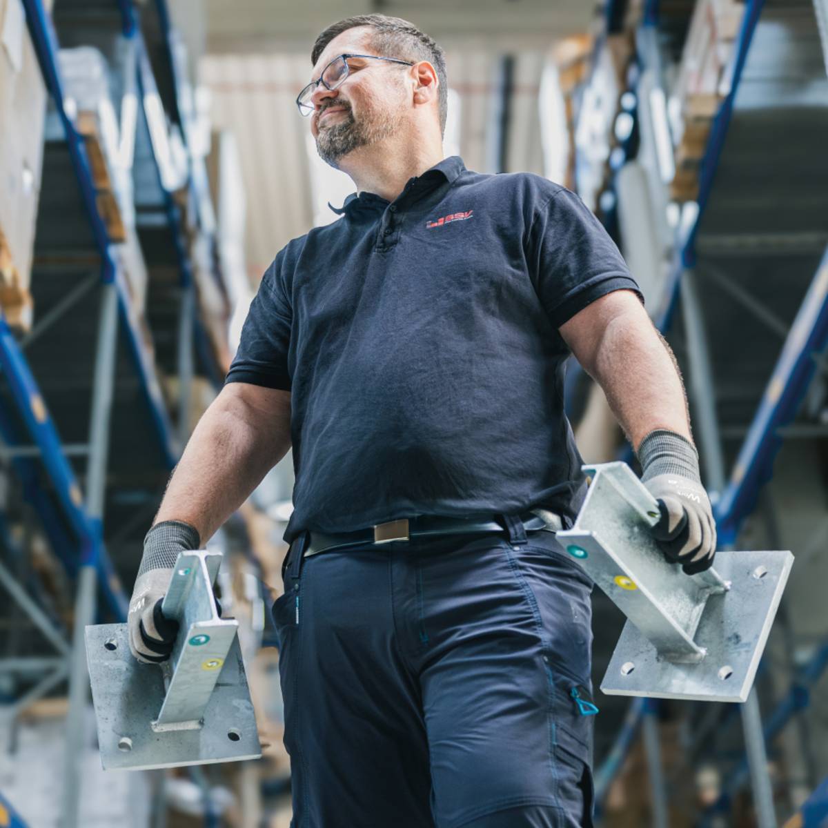 Eine Person in einer Lagerhausumgebung trägt eine dunkelblaue Uniform und hält zwei große Metallhalter in den Händen, während sie zwischen hohen Regalen mit gestapelten Waren steht.