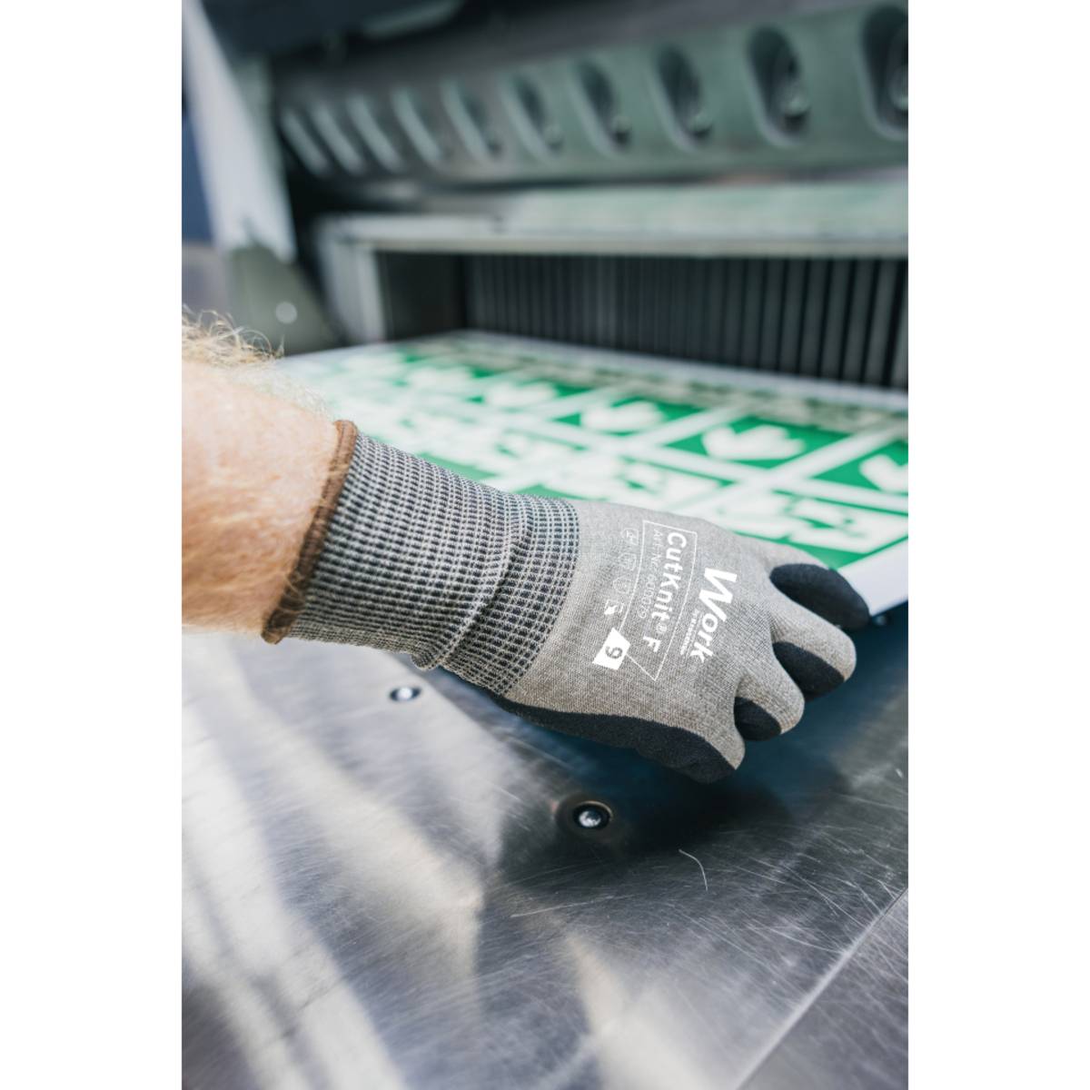 Eine behandschuhte Hand einer Person greift nach einem Stapel grün und weiß bedruckter Papiere, die von einer industriellen Papierschneidemaschine geschnitten werden.